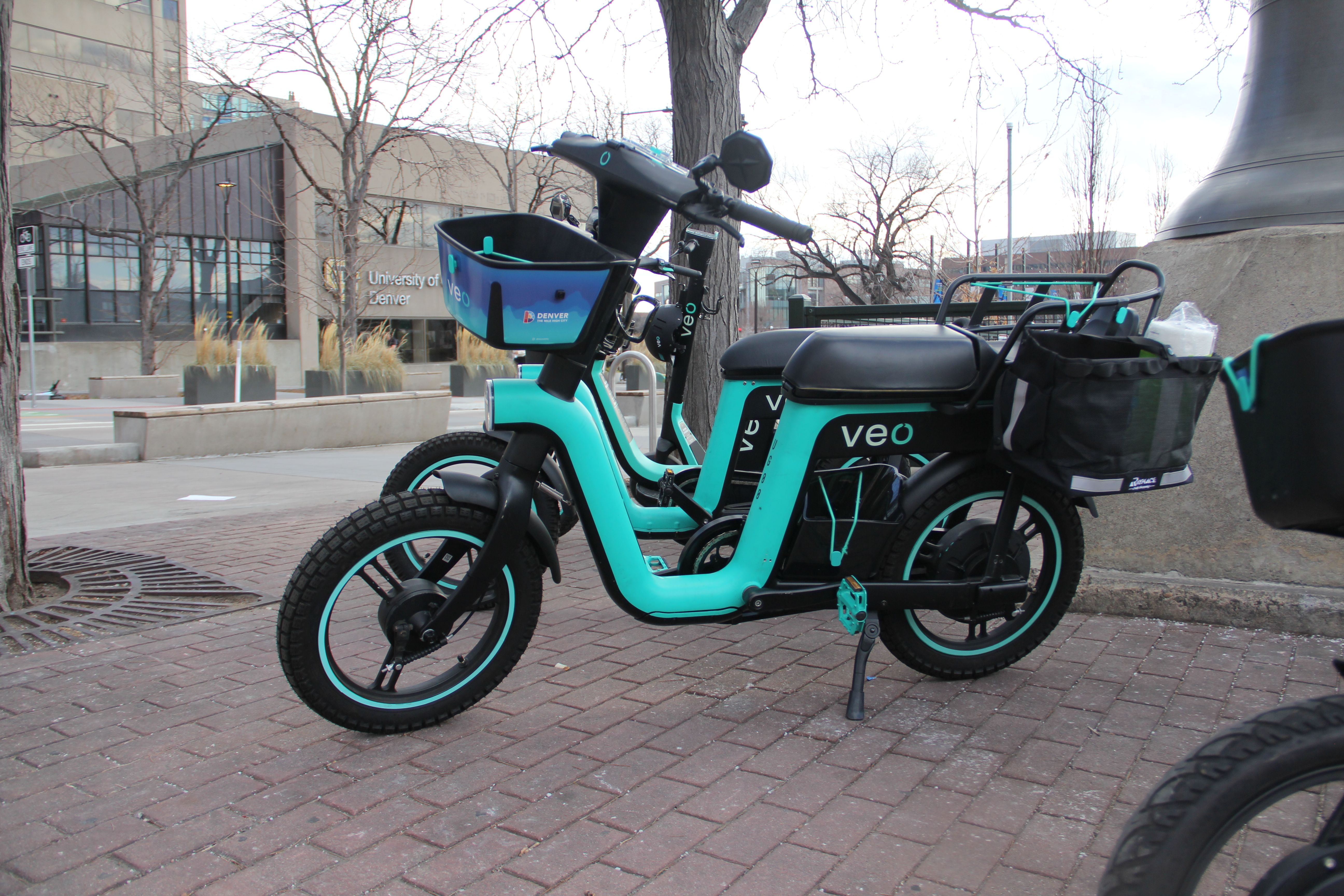 Two teal and black Veo electric bikes parked on a brick sidewalk near the University of Denver, with leafless trees and modern buildings in the background.