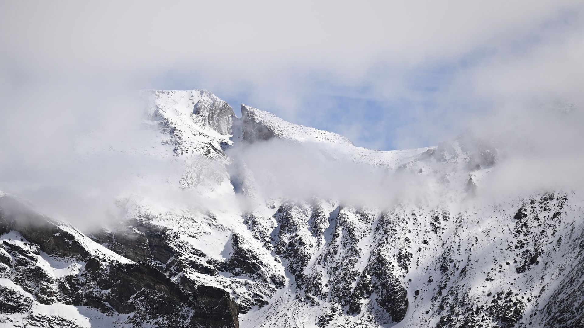 Snowy mountain ridge rises through low-lying clouds, with pale blue sky peeking through; rugged rock faces coated in snow along the lower slopes.