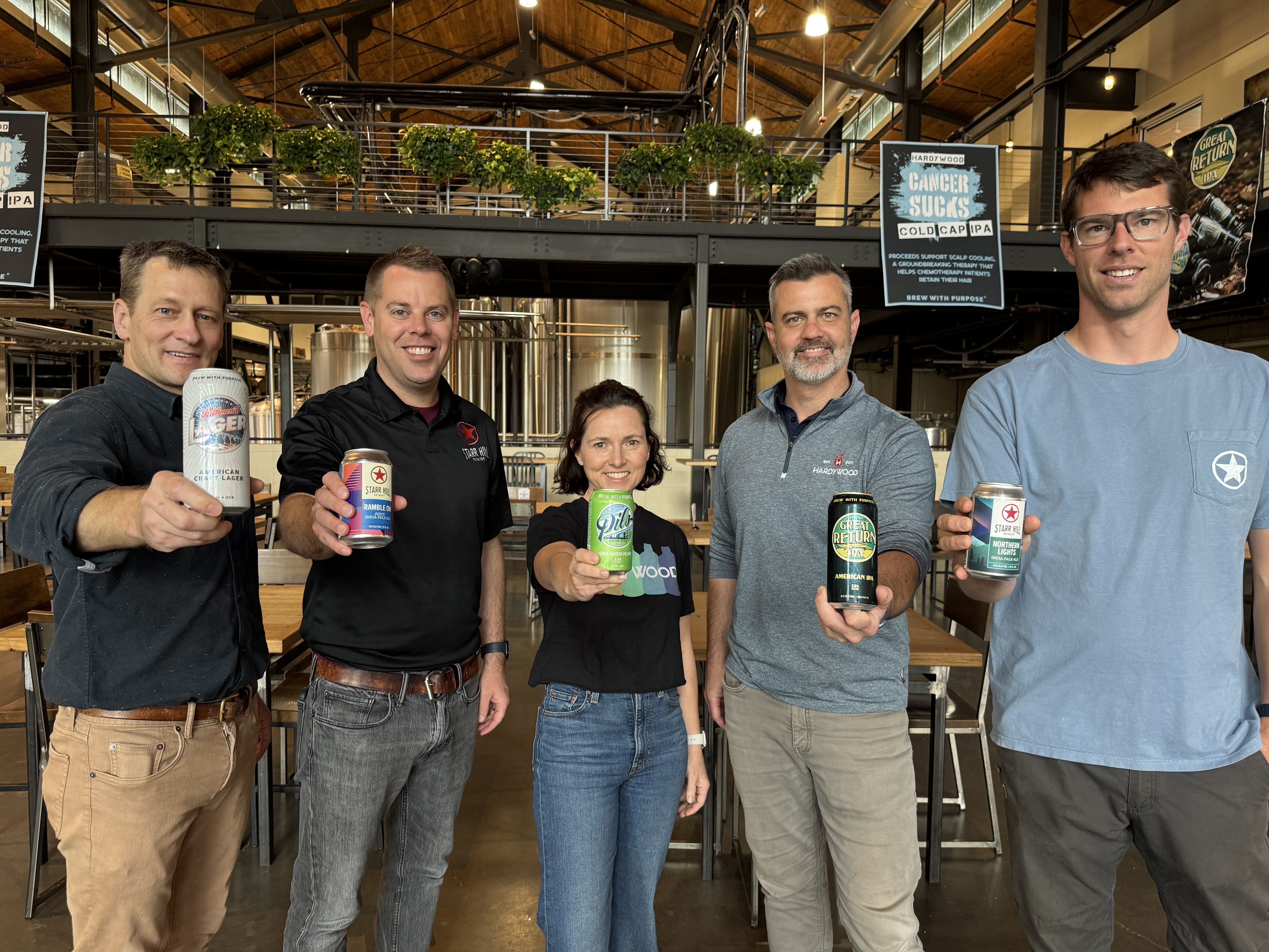 Five people stand in a brewery, smiling at the camera, each holding a beer can. Industrial space with exposed beams, plants, and posters in the background.