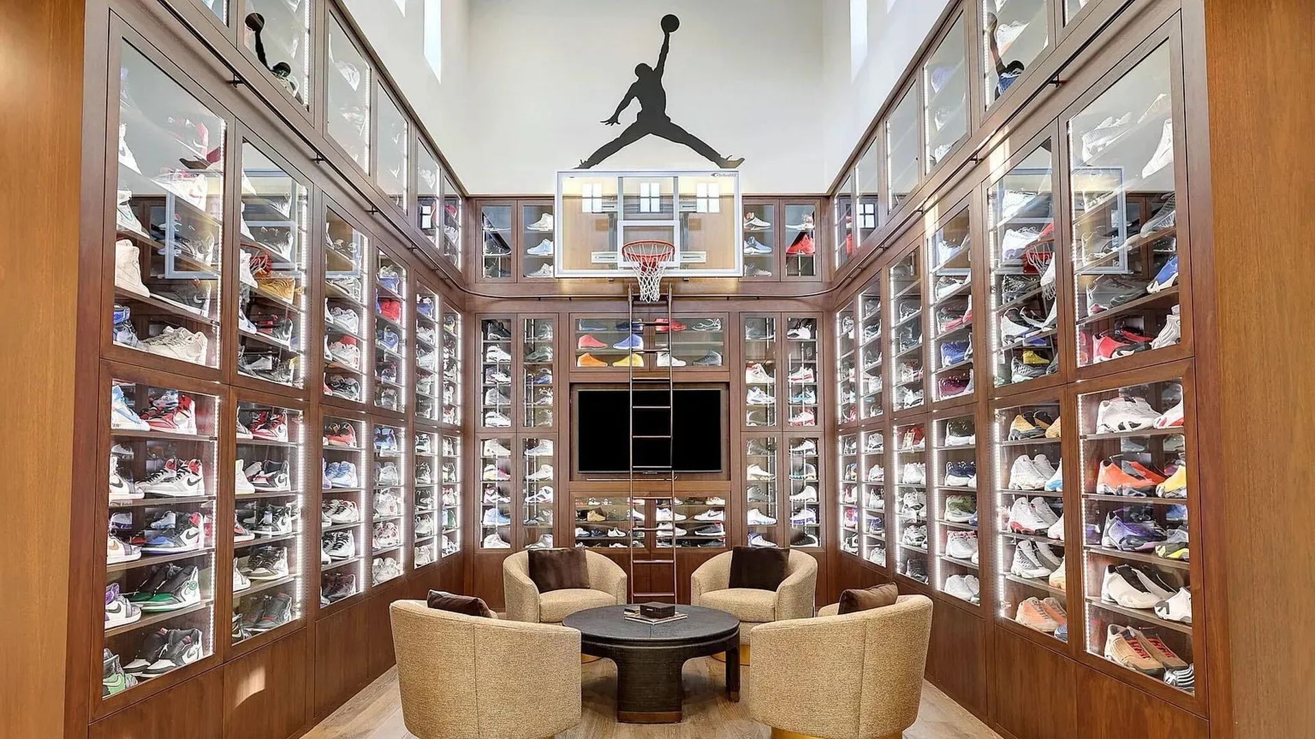 Room with wooden floors and walls covered in glass-fronted shelves filled with colorful sneakers. Four beige armchairs surround a black round table. Basketball hoop and Jumpman logo above TV.
