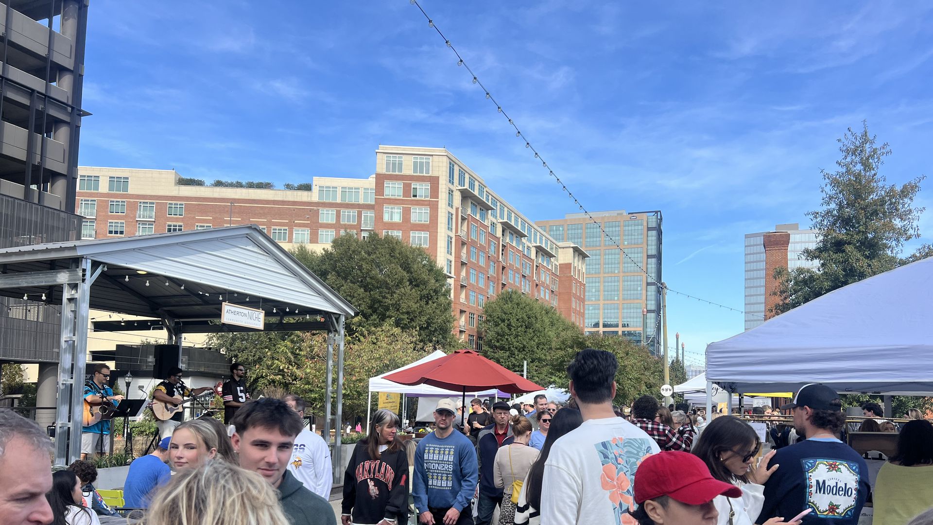 Crowd at an outdoor market with people walking and socializing around stalls and food stands, musicians playing on a small stage, under a bright blue sky and surrounded by tall buildings.