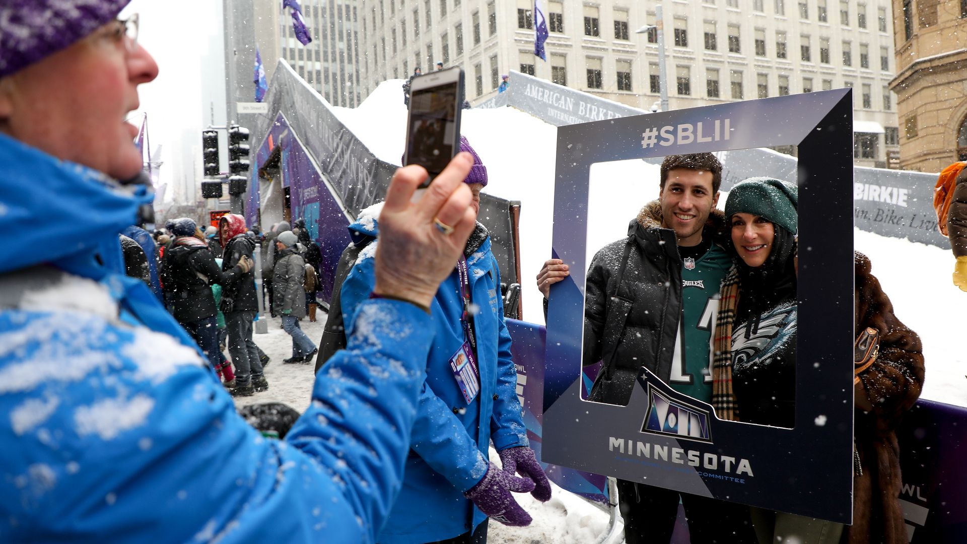 People in winter clothes posing outdoors in snow, holding a large photo frame with #SBLII and Minnesota logo, while another person in blue jacket takes their picture on a smartphone.