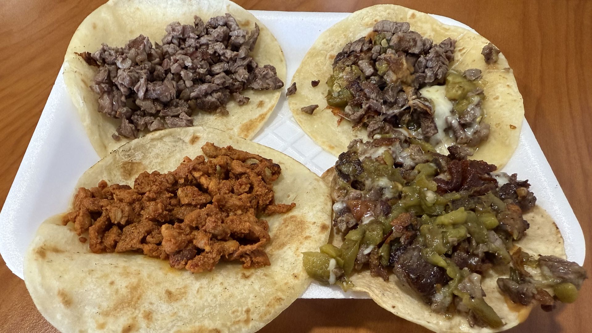 Four open-faced soft tacos on a white foam tray, two with chopped grilled steak, one with pastor pork, and one with steak, melted cheese, and green peppers.