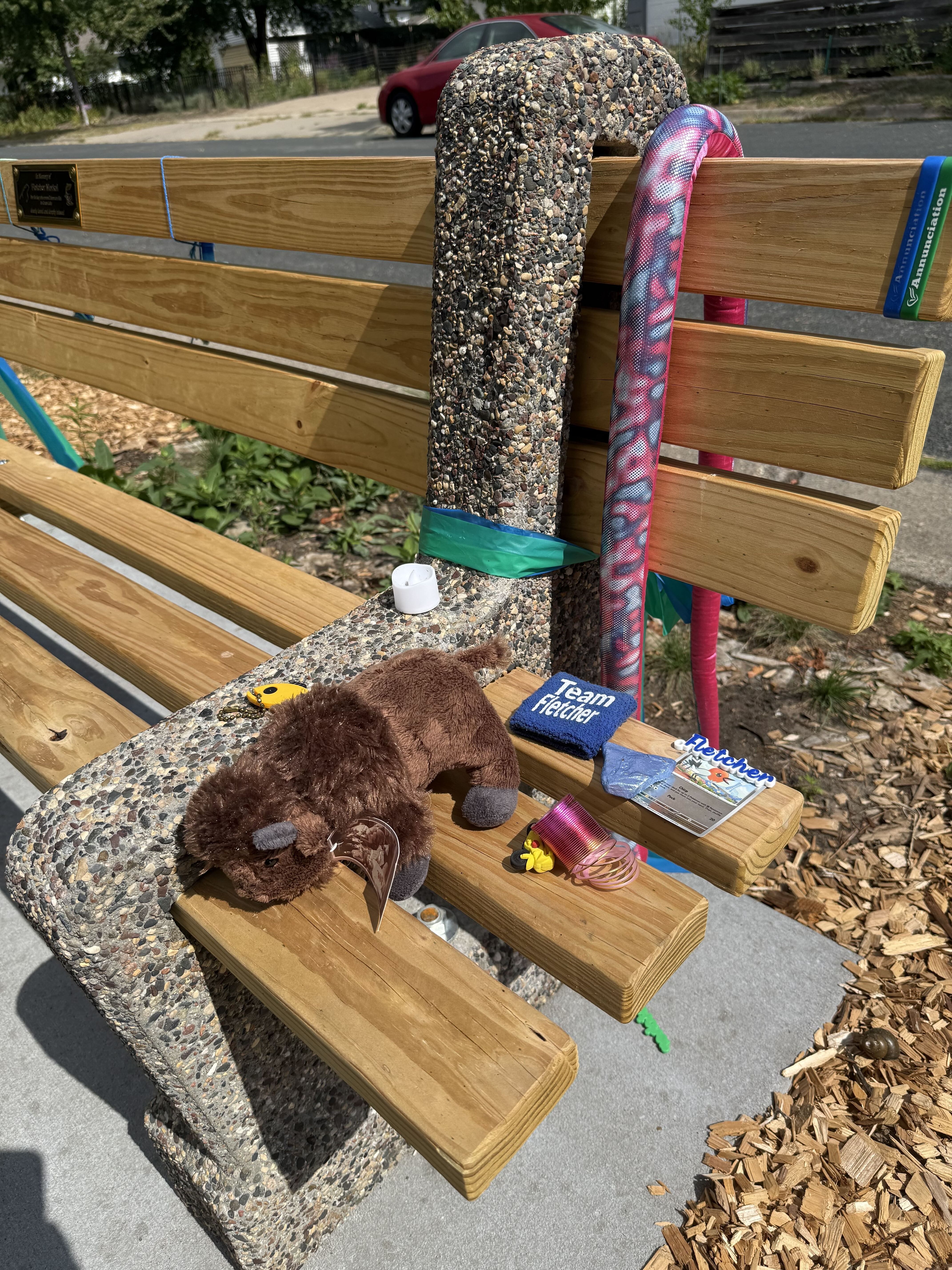 Wooden park bench with a pebble-textured curved post wrapped in teal tape. Pink-blue patterned pool noodle drapes over the back; a brown plush toy, Team Fighter patch, and a magazine on the seat.