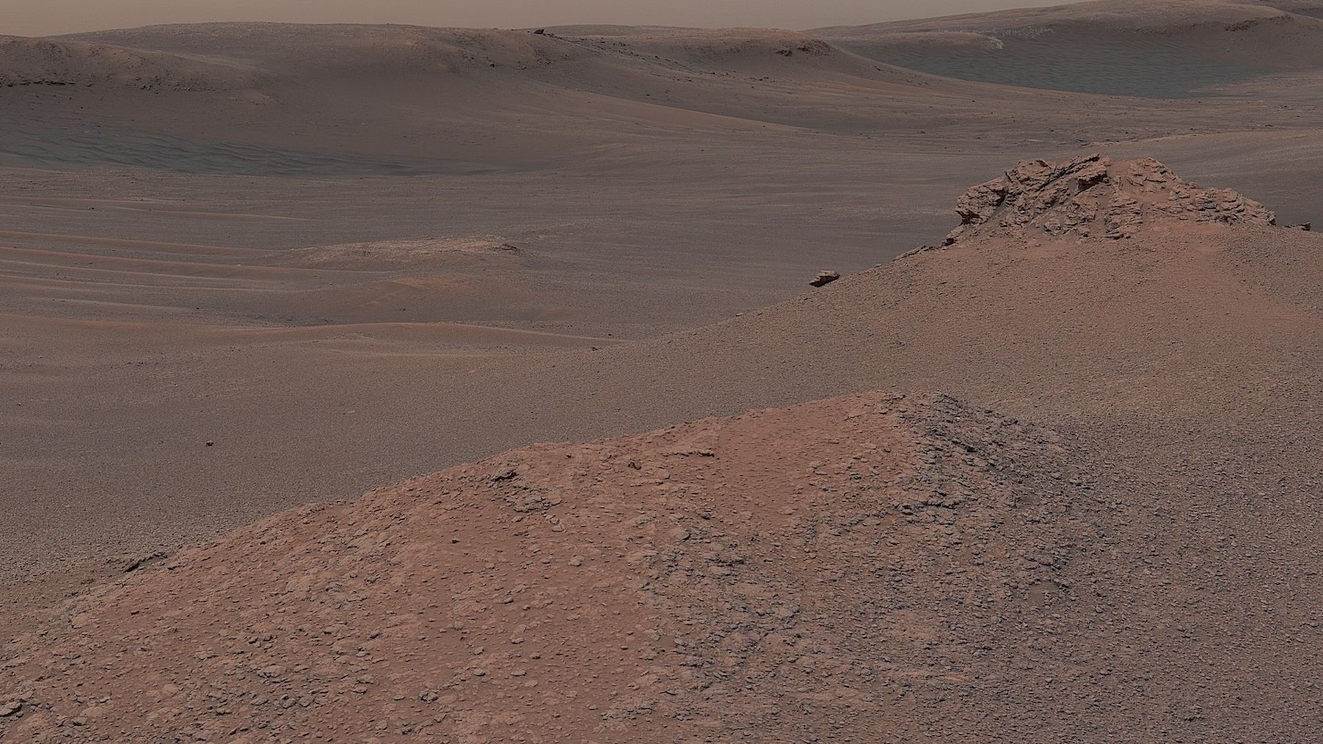 A landscape on Mars in red with a rocky outcropping on the right and waves of sand on the left