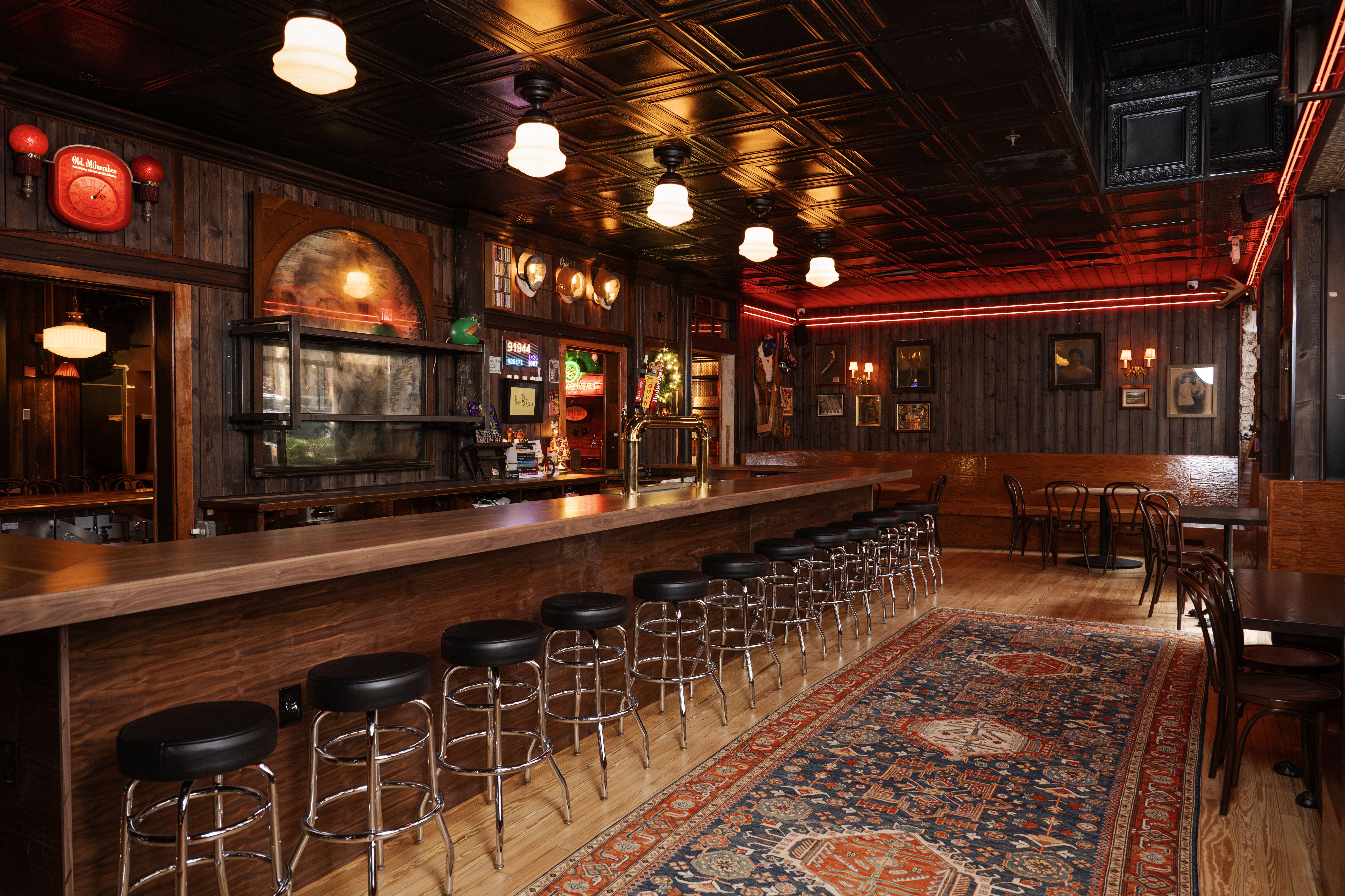 Empty cozy bar with wooden walls, a long wooden bar counter with black stools, vintage decor, warm ceiling lights, neon red strip lighting, and a patterned rug on the wooden floor.