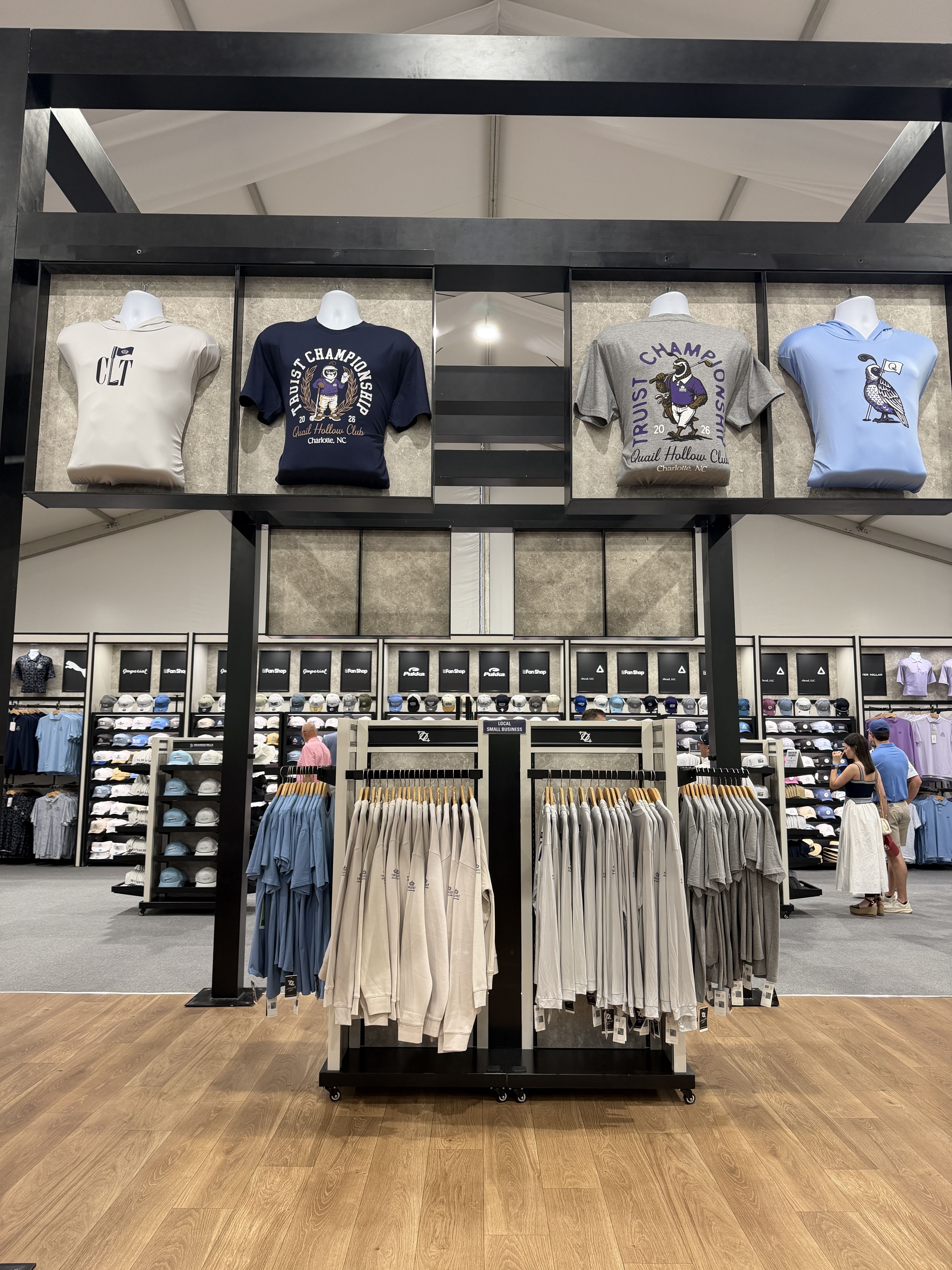 Inside a clothing store, four large tee displays on a black metal frame above racks of shirts. Beige, navy, gray, and light-blue graphic tees with sports designs; wood floor.