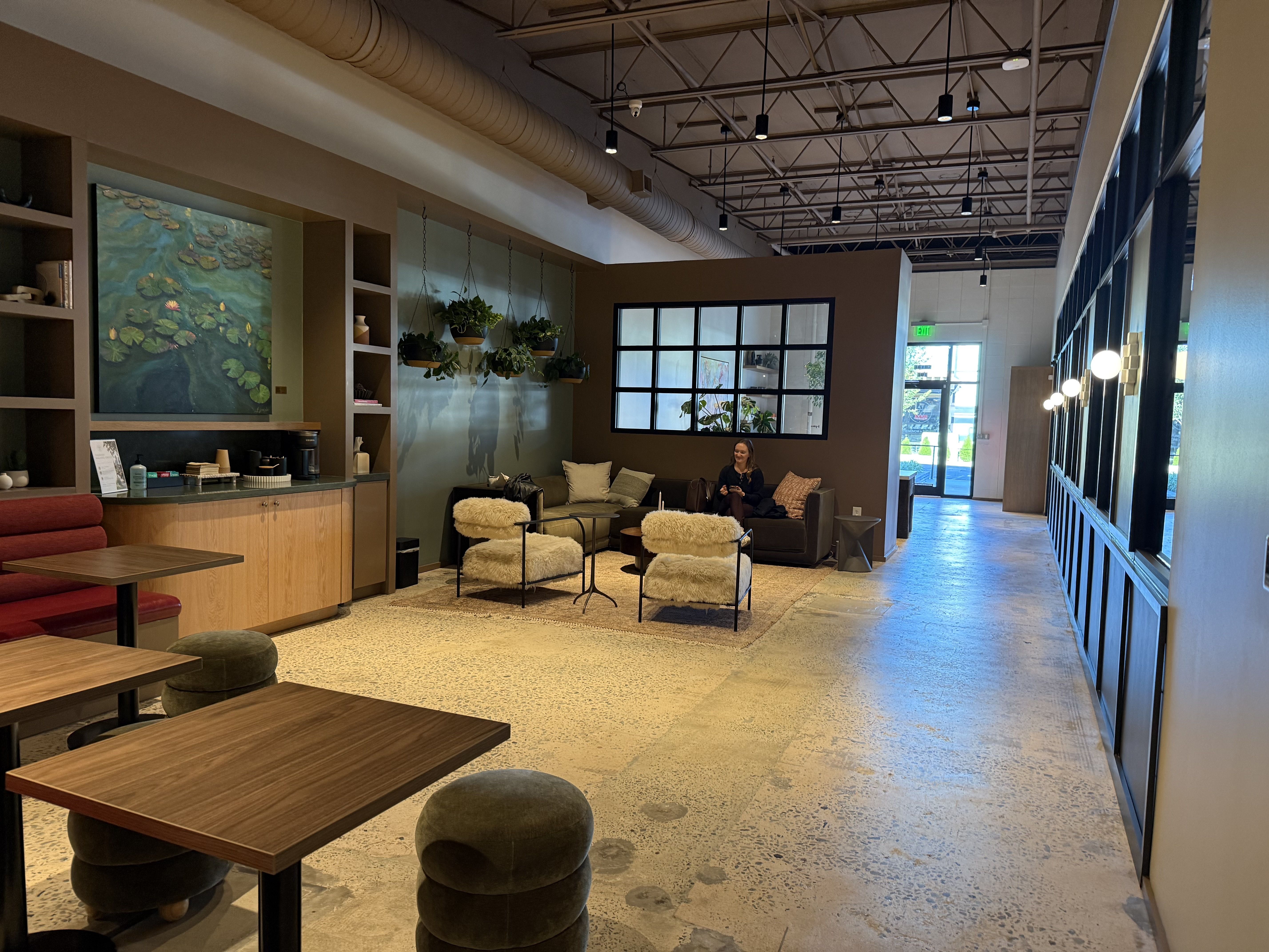 Interior of a modern cafe lobby with a glass wall on the right, hanging plants, and a water-lily mural. Cozy seating with fluffy ottomans; a woman sits on a sofa. Exposed ceiling beams and pendant lights over terrazzo flooring.