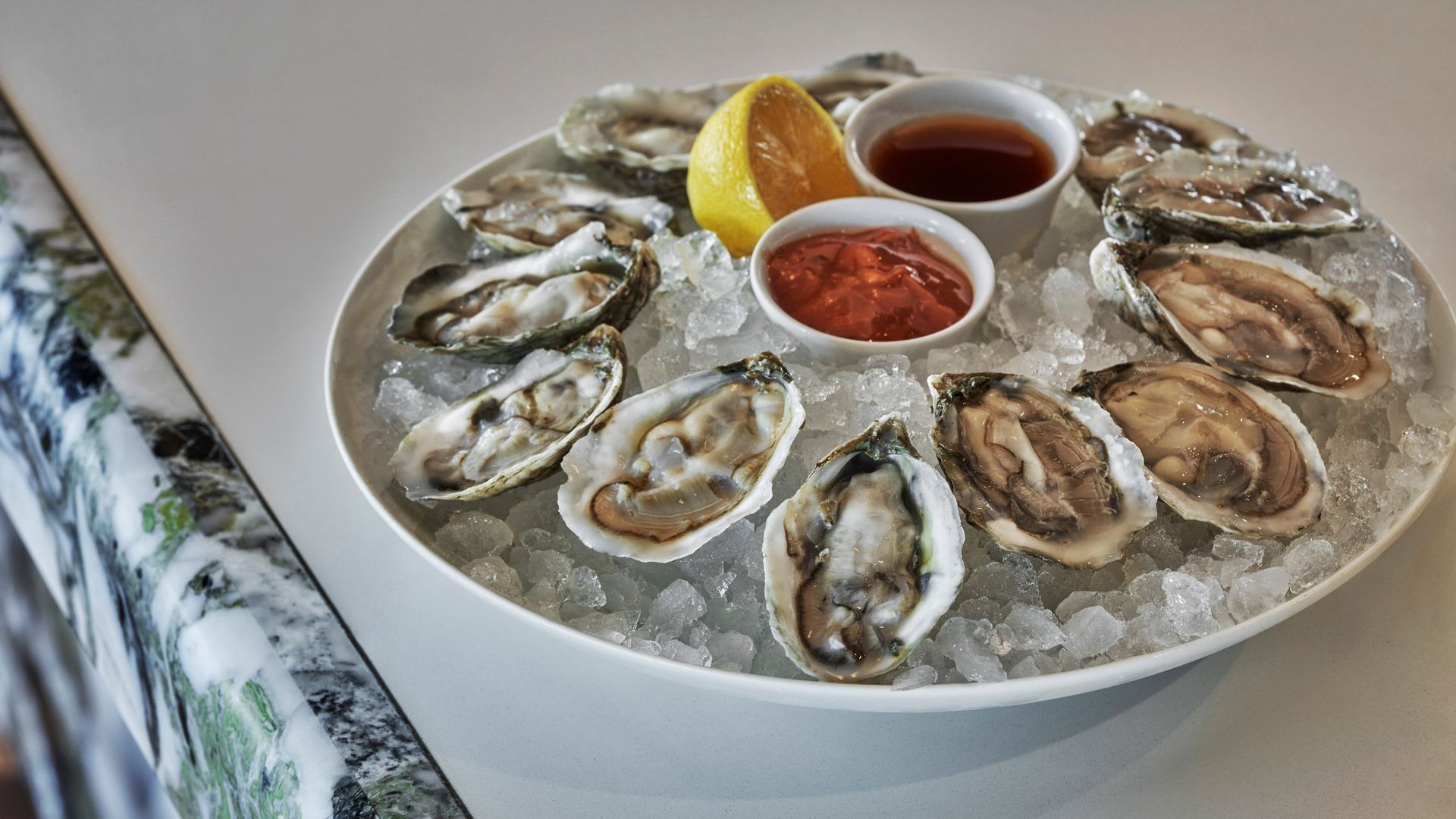 A platter of oysters on the half-shell sits on a bartop. The platter is filled with ice, oysters, two sauces and a lemon wedge.
