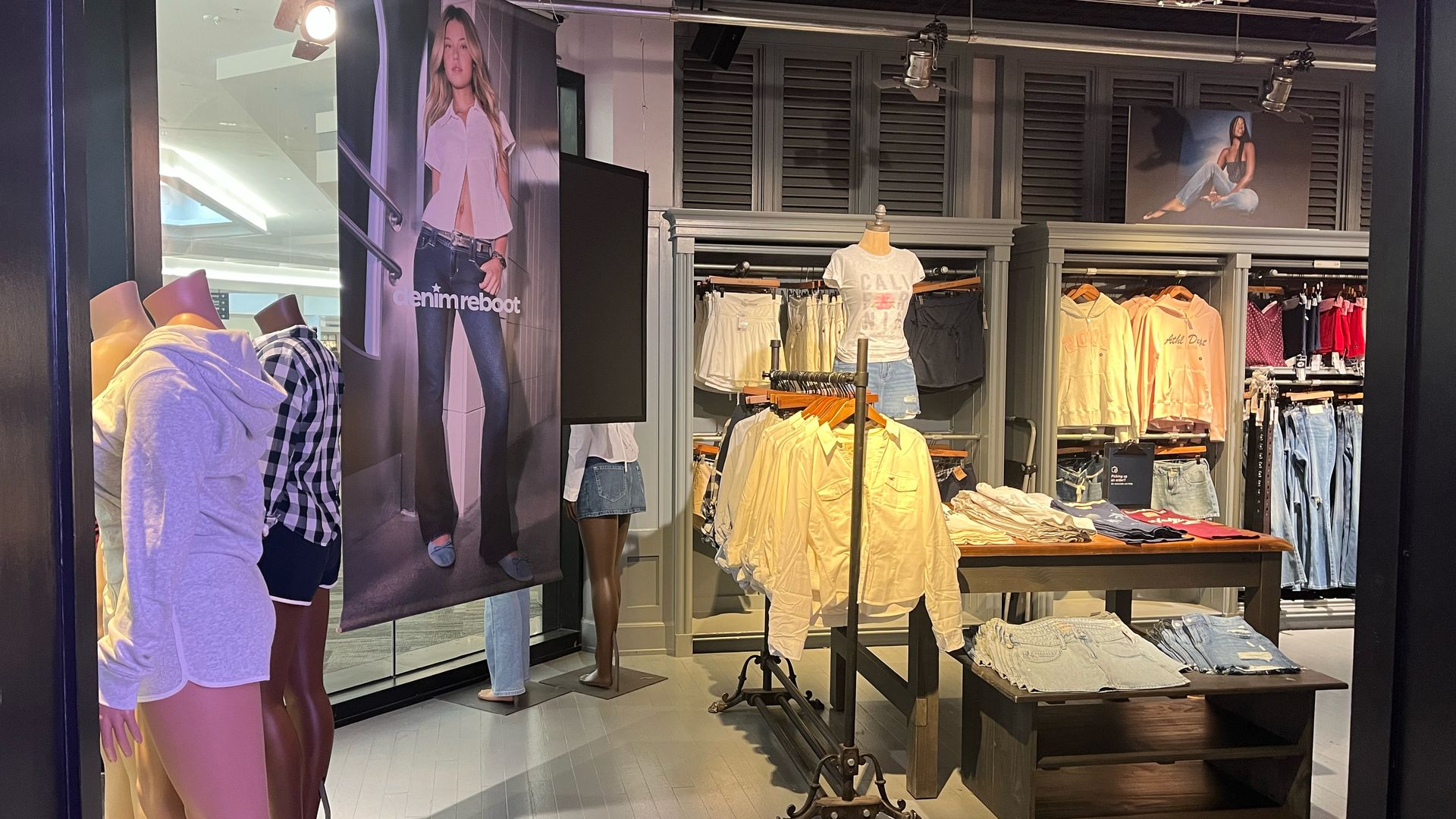 Hollister clothing store display with mannequins dressed in casual outfits, racks of button-up shirts, denim shorts, and hoodies in pastel colors, and posters of models wearing jeans.