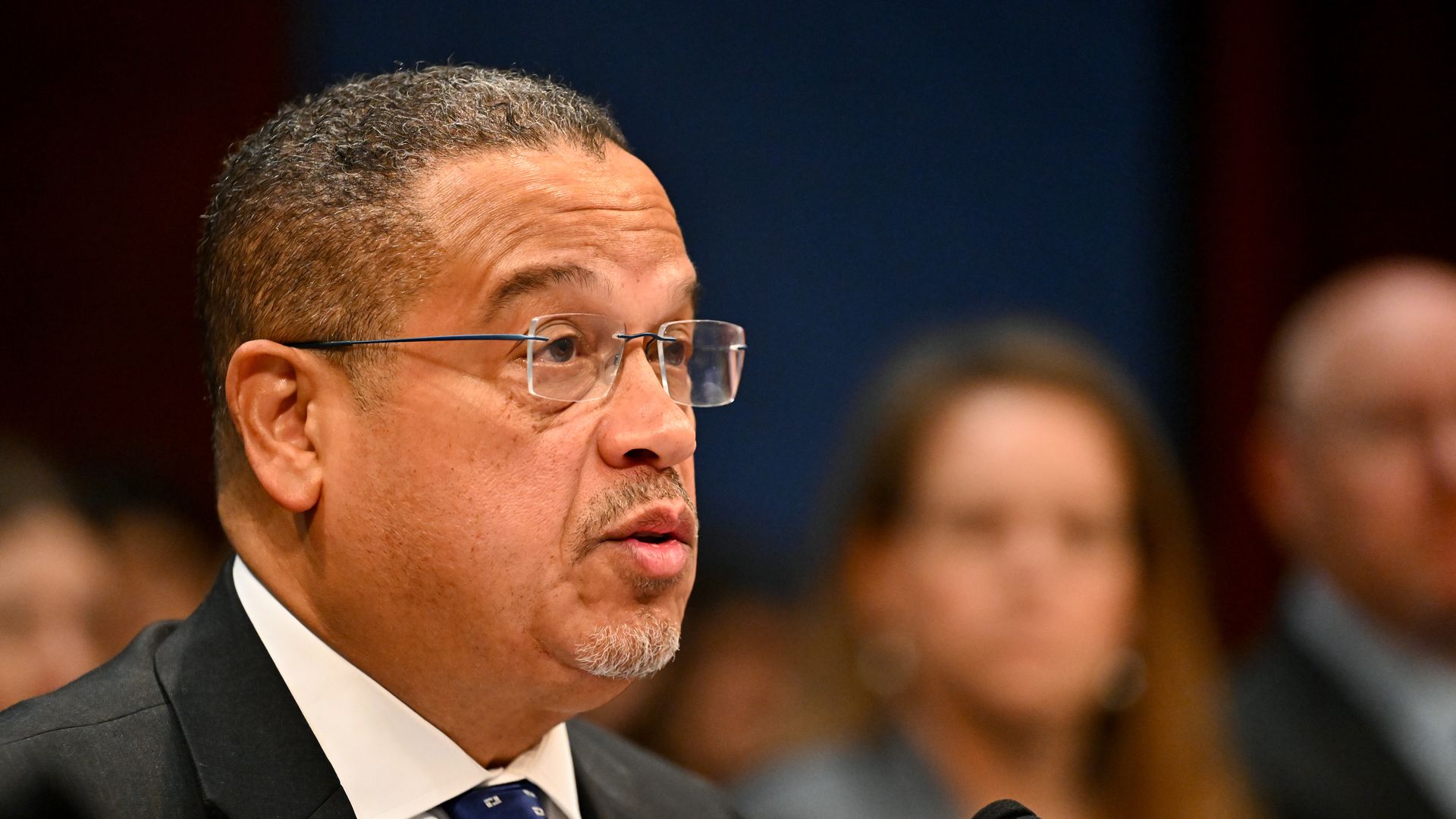 Close-up of a Keith Ellison in a dark suit and glasses, speaking into a microphone; blue tie and lapel pin visible, blurred audience in the background.
