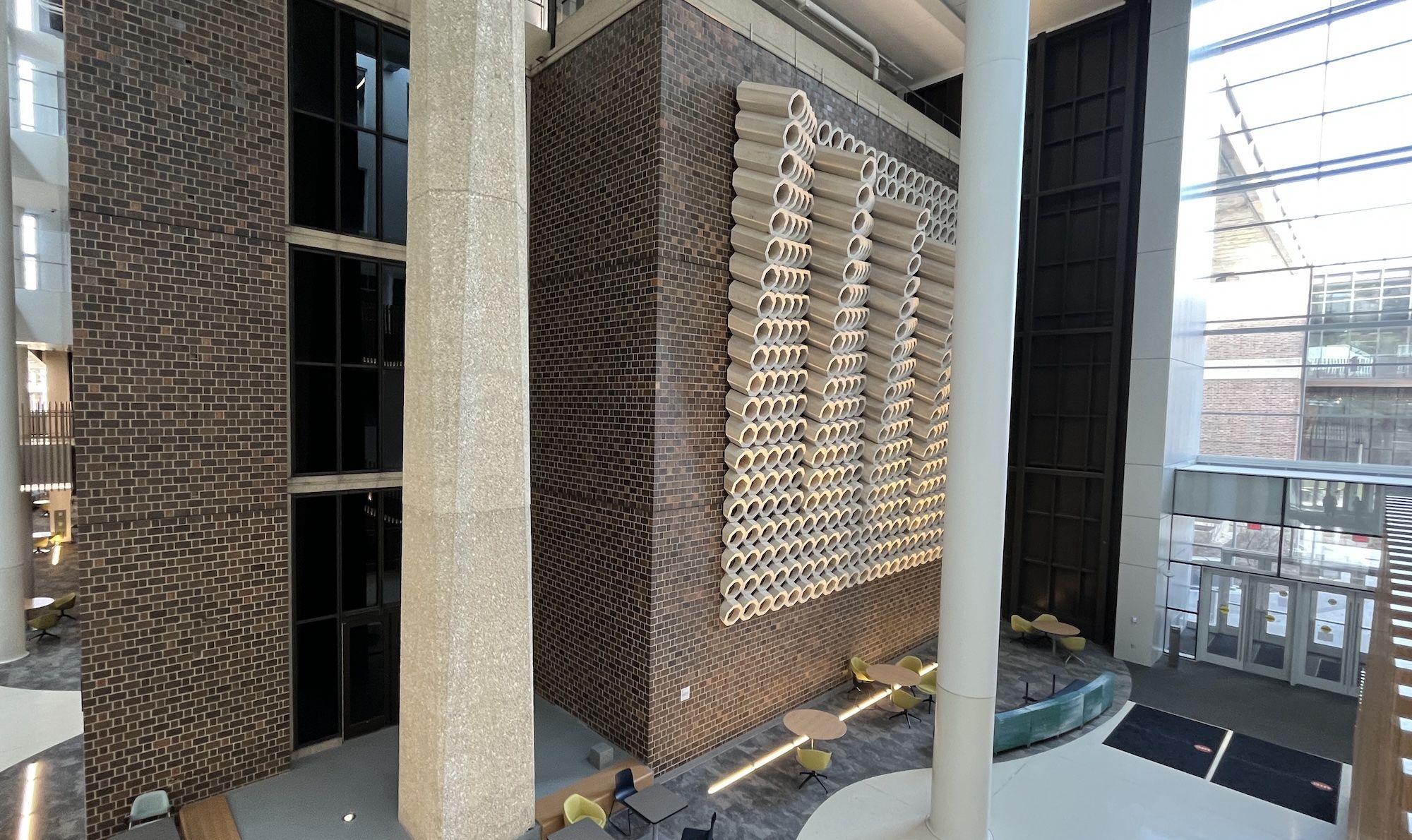 Atrium interior with brown brick walls and a wall sculpture of stacked white tubes. A tall beige pillar and a white column stand beside yellow seating and glass doors.