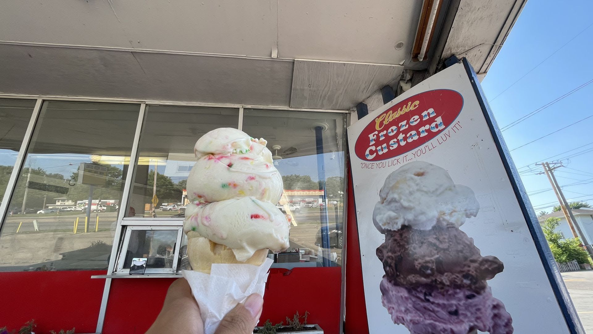 Three scoops of birthday cake ice cream on a cone held up next to a Classic Frozen Custard sign.