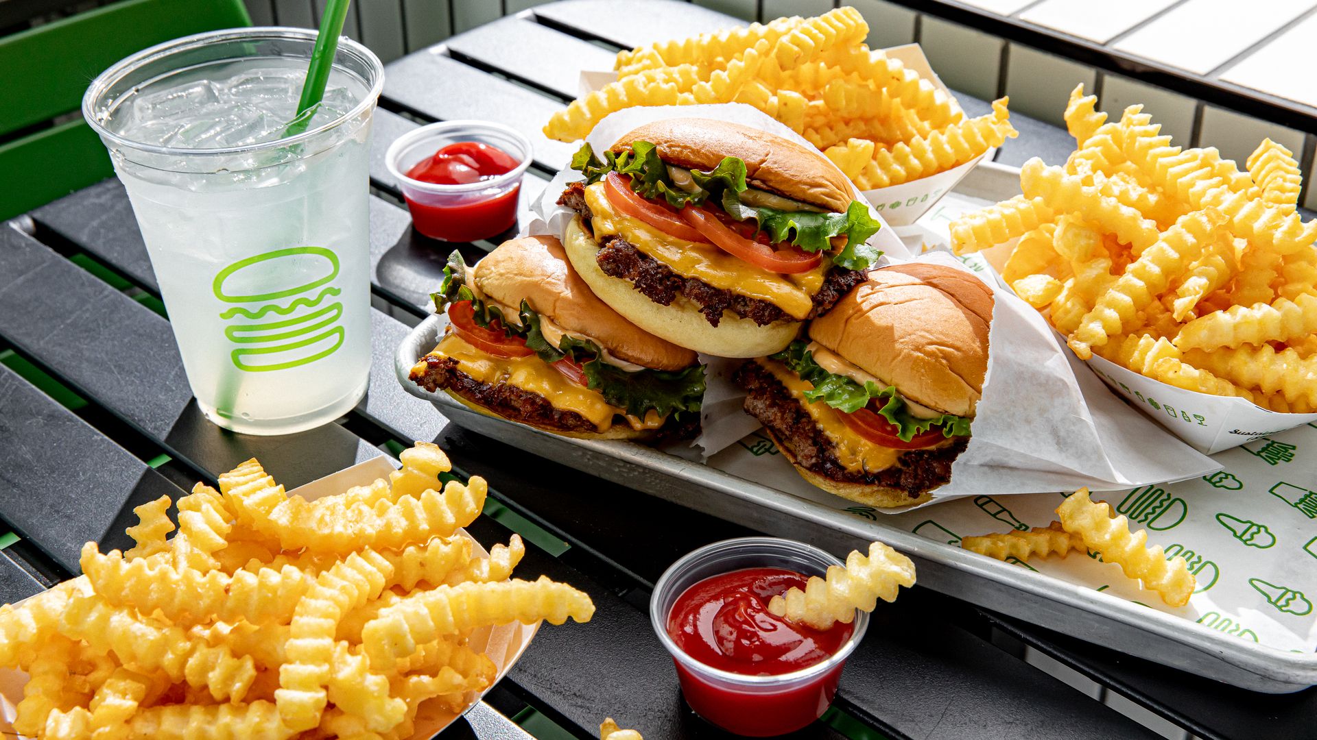 Two cheeseburgers with lettuce, tomato, pickles and melted cheese on buns, a large iced drink with a green straw, and abundant crinkle-cut fries with ketchup on a metal table.