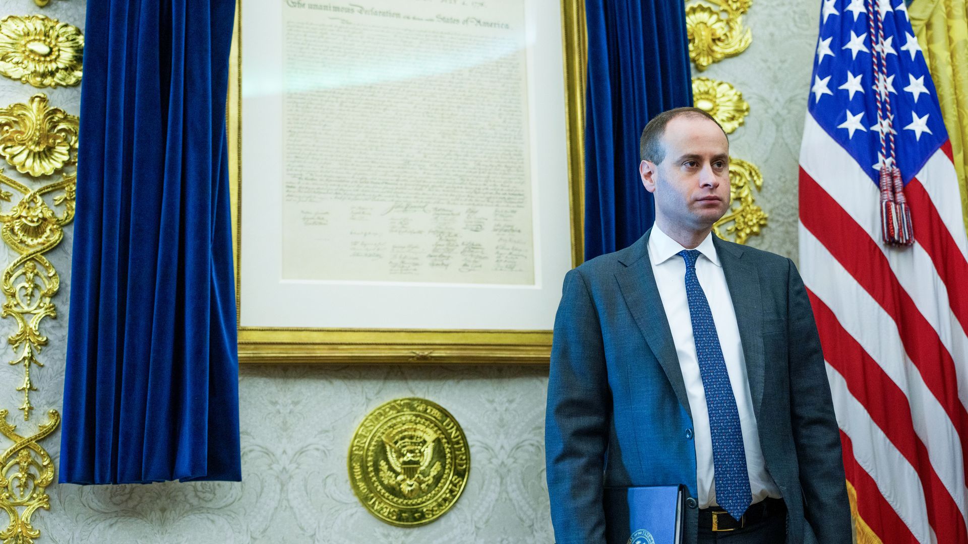 White House staff secretary Will Scharf stands in the Oval Office beside a U.S. flag, with a framed copy of the Declaration of Independence behind him.