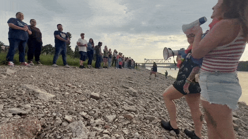 A diverse group of people standing in a long line on a rocky riverbank at sunset, with two women in the foreground, one speaking into a megaphone.