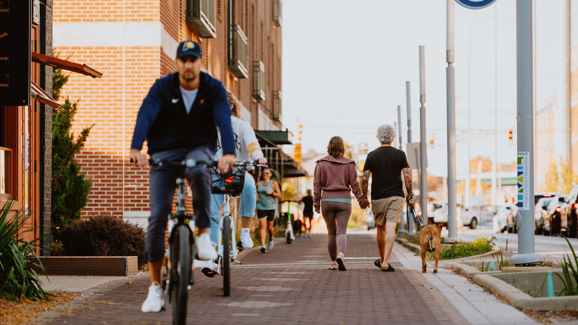 Urban street scene along a brick sidewalk: cyclists and pedestrians, a man in a blue jacket rides toward camera, a couple walks a dog, and a round sign reads "Cultural Trail" on the right.