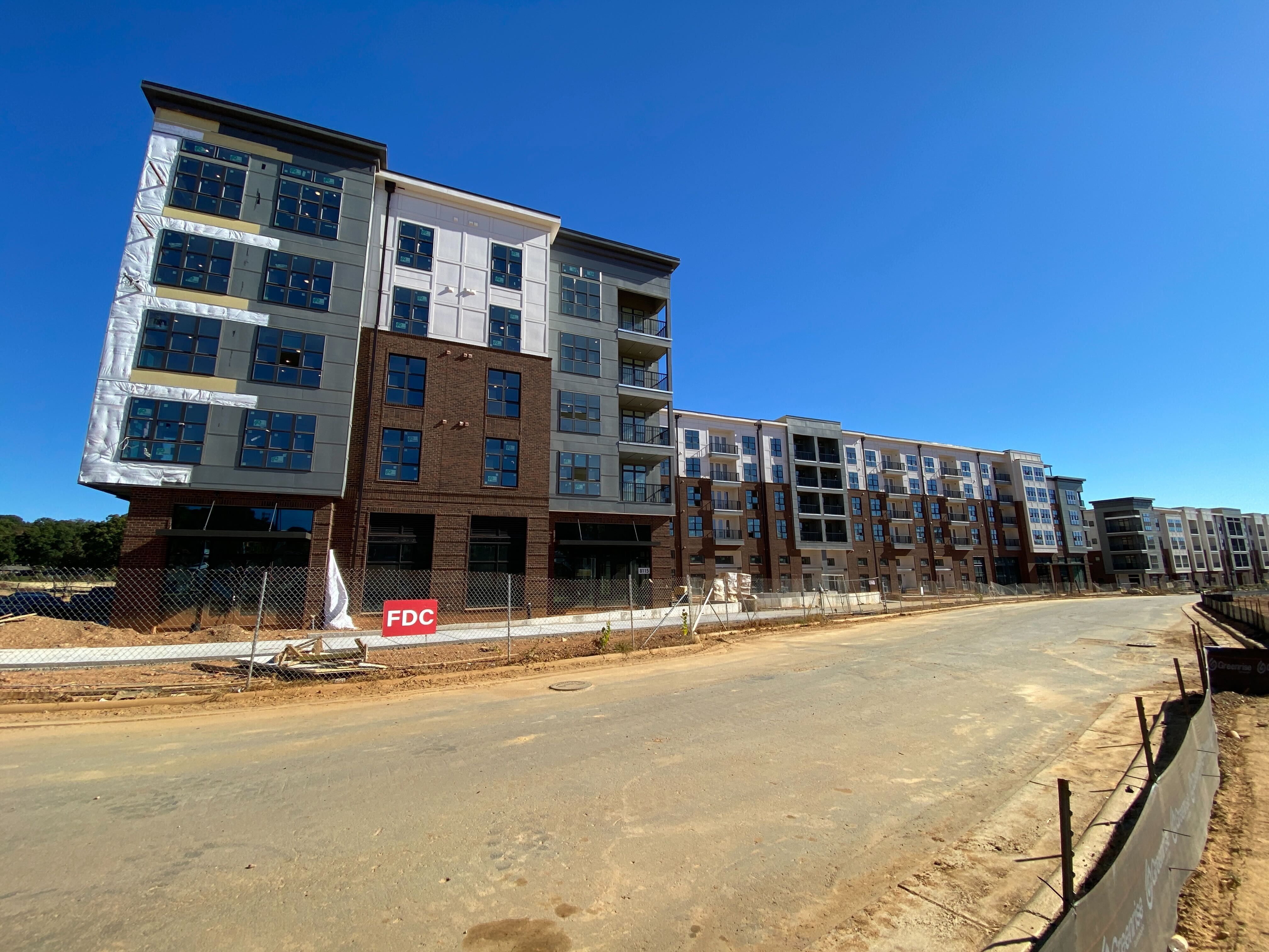 Large multi-story apartment building under construction with brown and gray exteriors, many windows, balconies, fenced-off dirt road, clear blue sky overhead.