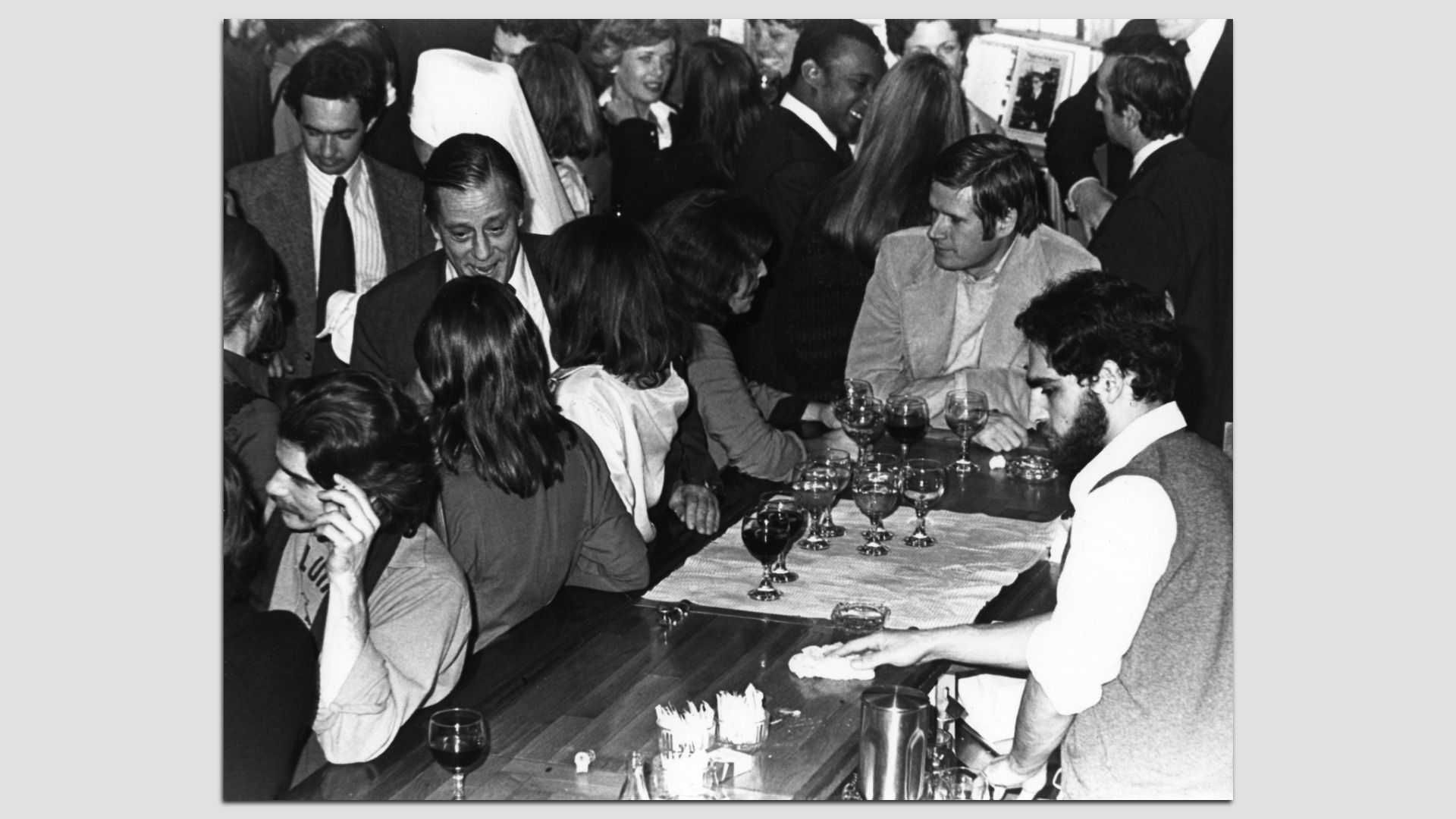 Black-and-white photo of a crowded bar scene, people gathered around a long counter with wine glasses; a bearded bartender in a vest stands on the right and a waiter is in the foreground.