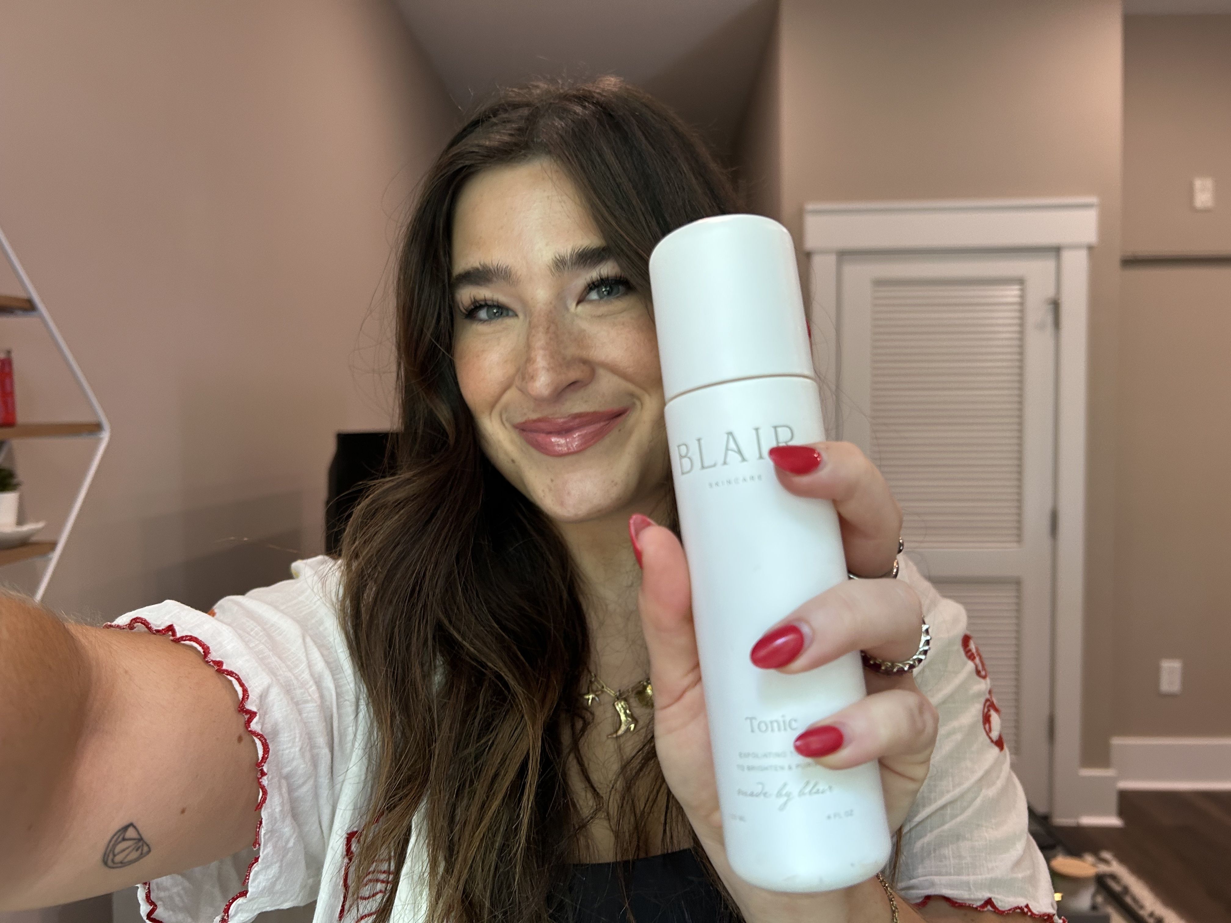 Smiling woman with long brown hair and red nails holding a white bottle labeled "BLAIR Skincare Tonic" indoors with neutral walls and a door in the background.
