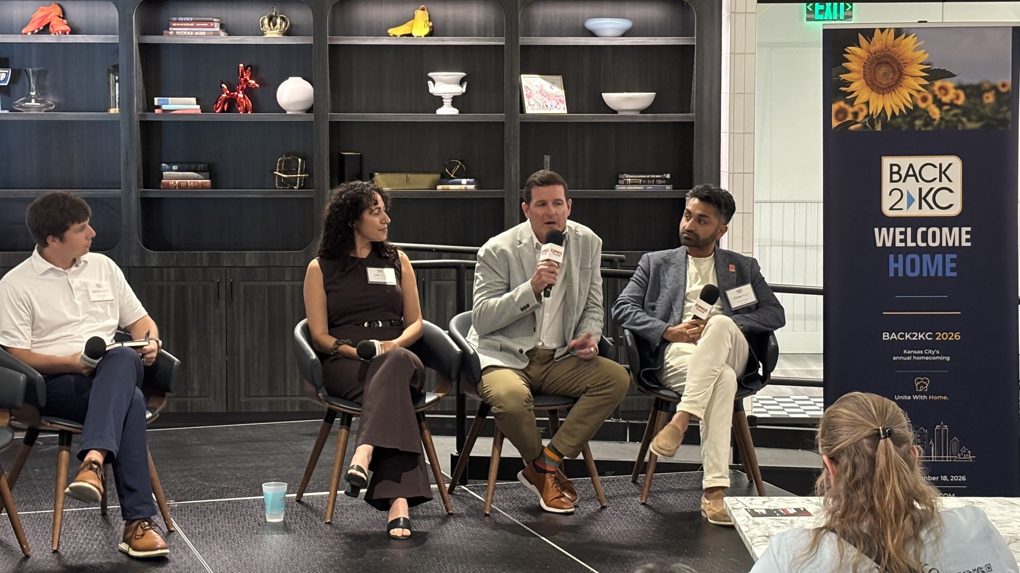 Panel of four speakers seated on a stage in front of dark shelving; each holds a microphone. A large banner on the right says BACK 2 KC WELCOME HOME with sunflowers; audience visible in foreground.