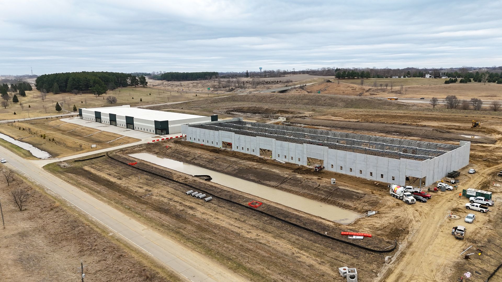 Drone view of a construction site with two long gray warehouse buildings under construction, muddy trenches, pipes, and vehicles along a dirt road amid open fields under an overcast sky.