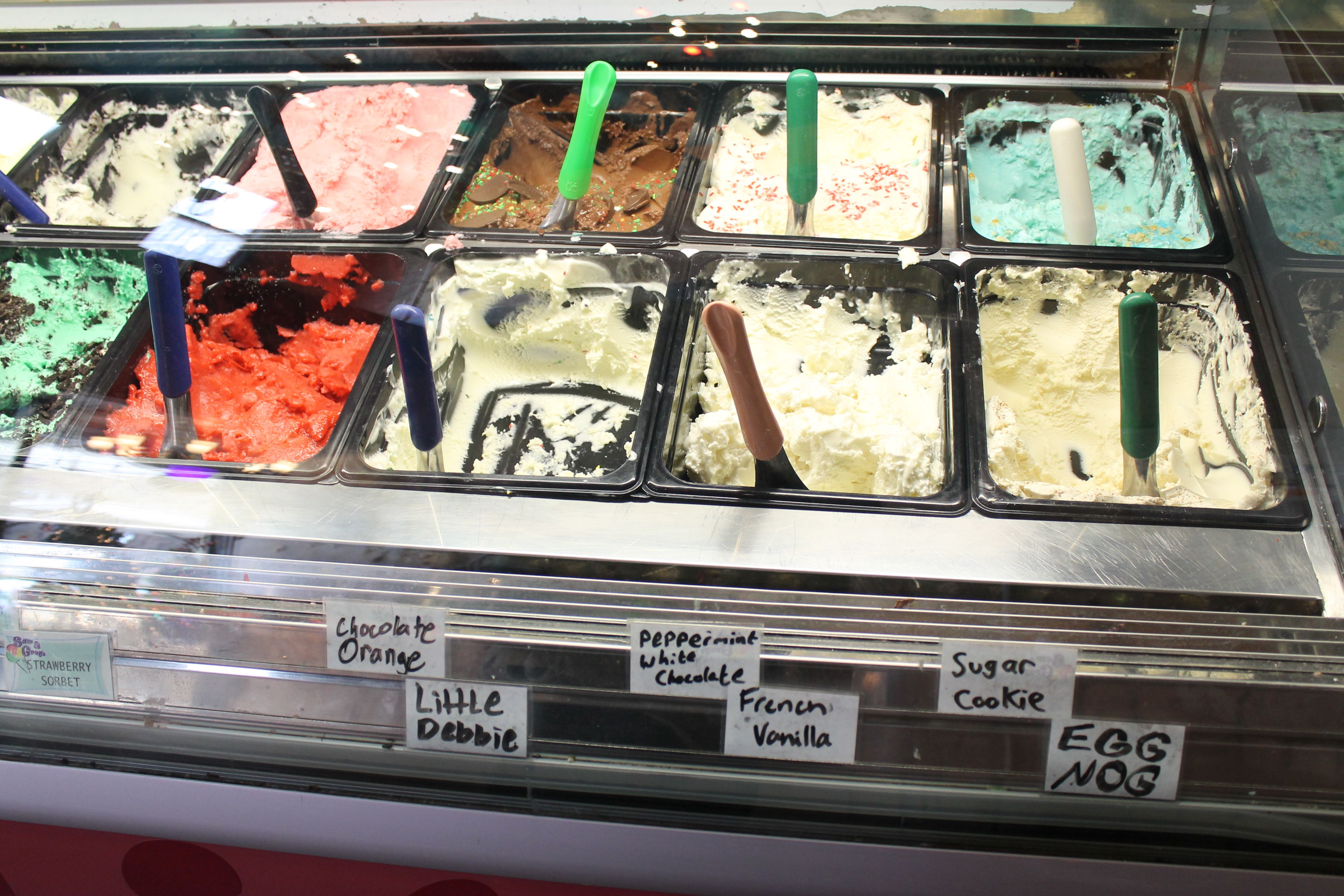 Ice cream display with various flavors including strawberry sorbet, chocolate orange, Little Debbie, peppermint white chocolate, French vanilla, sugar cookie, and egg nog. Colorful scoops visible.