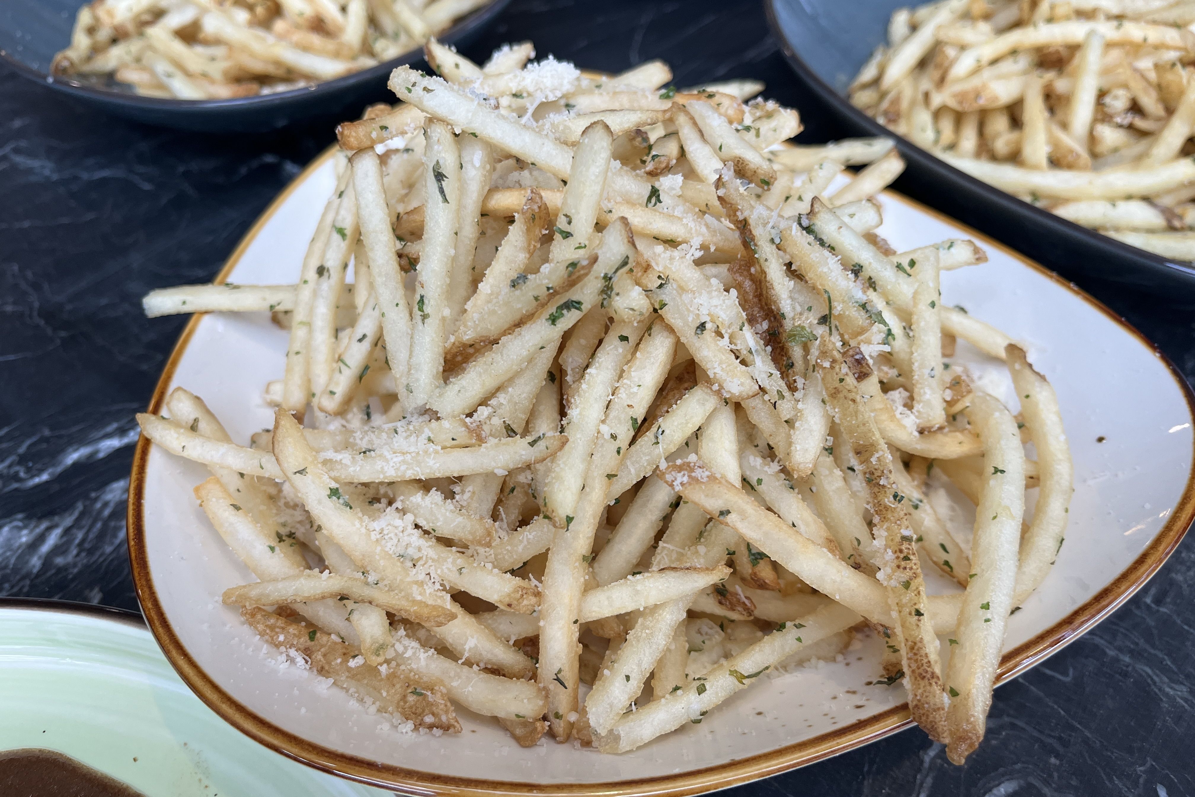 A mound of thin, crispy French fries sprinkled with grated cheese and chopped parsley on a white plate with a brown rim, set on a dark marble table.