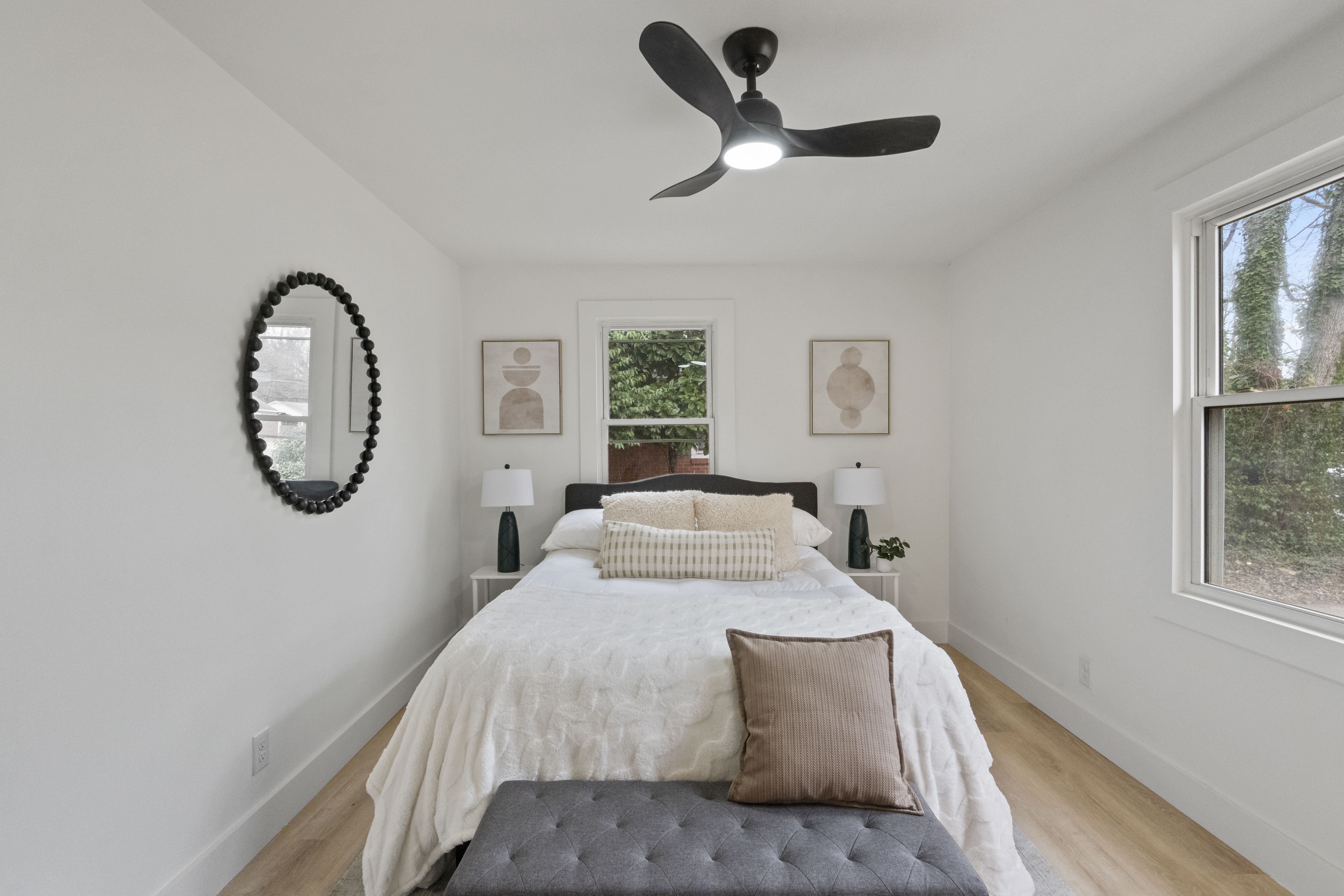 Minimalist bedroom with white walls, a bed with white bedding and beige pillows, a gray tufted bench at the foot, black ceiling fan, two bedside lamps, round mirror, and abstract art by window.
