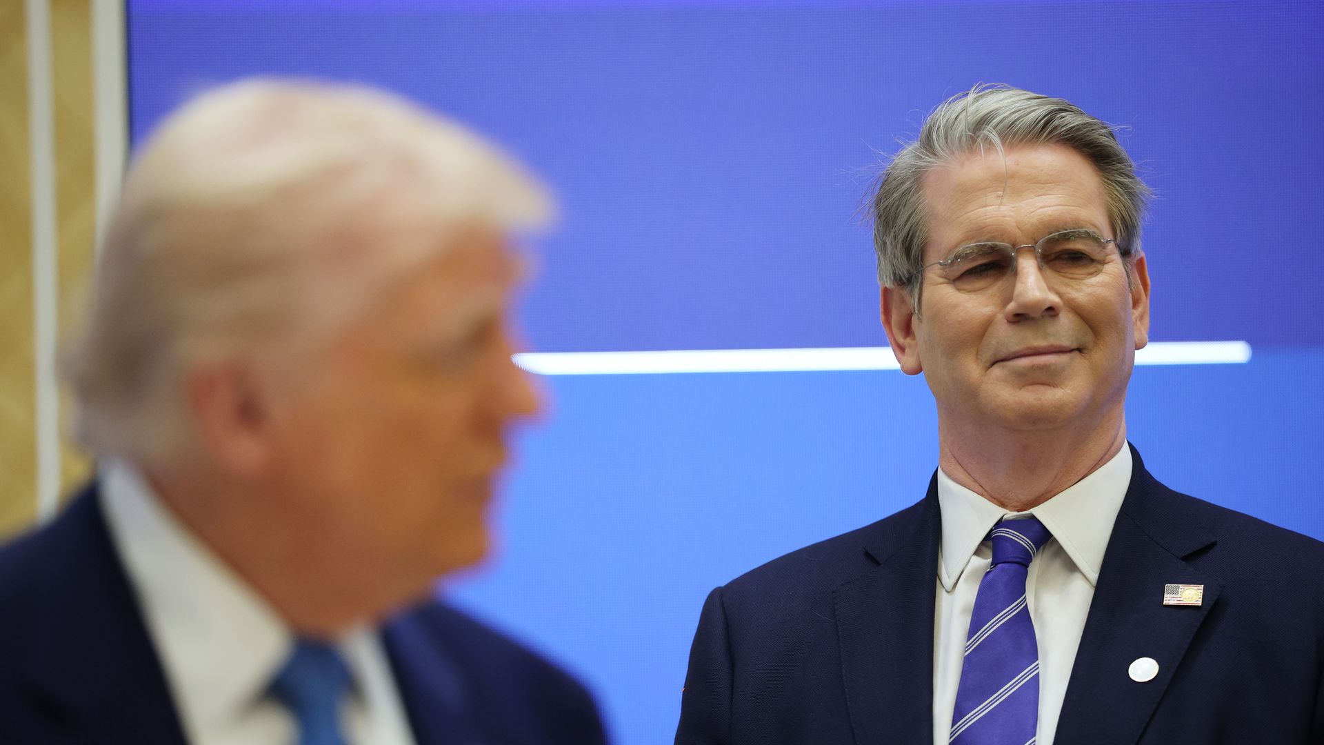 Treasury Secretary Scott Bessent, in the background, smiles as President Trump speaks.