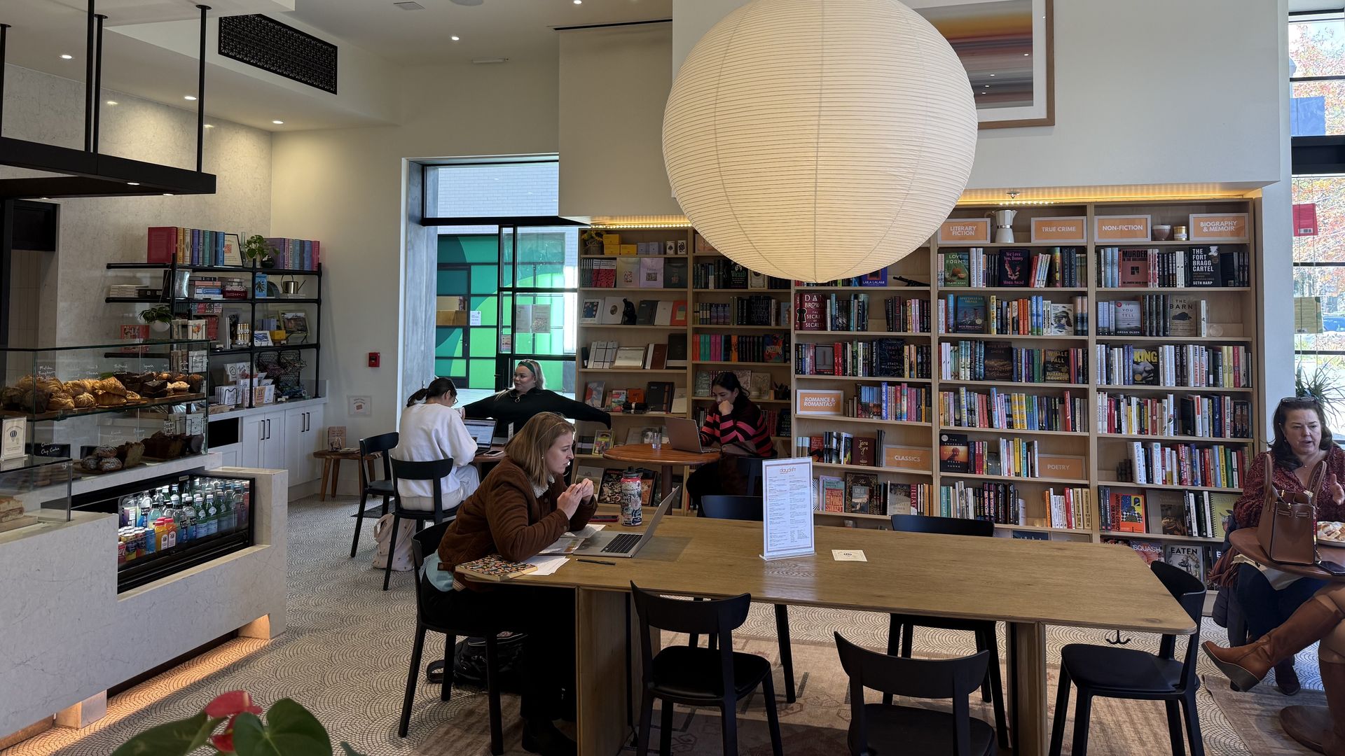 Bright bookstore cafe with wooden tables, black chairs, a large round paper lantern, bookshelves labeled by genre, and people reading, working on laptops, and eating.