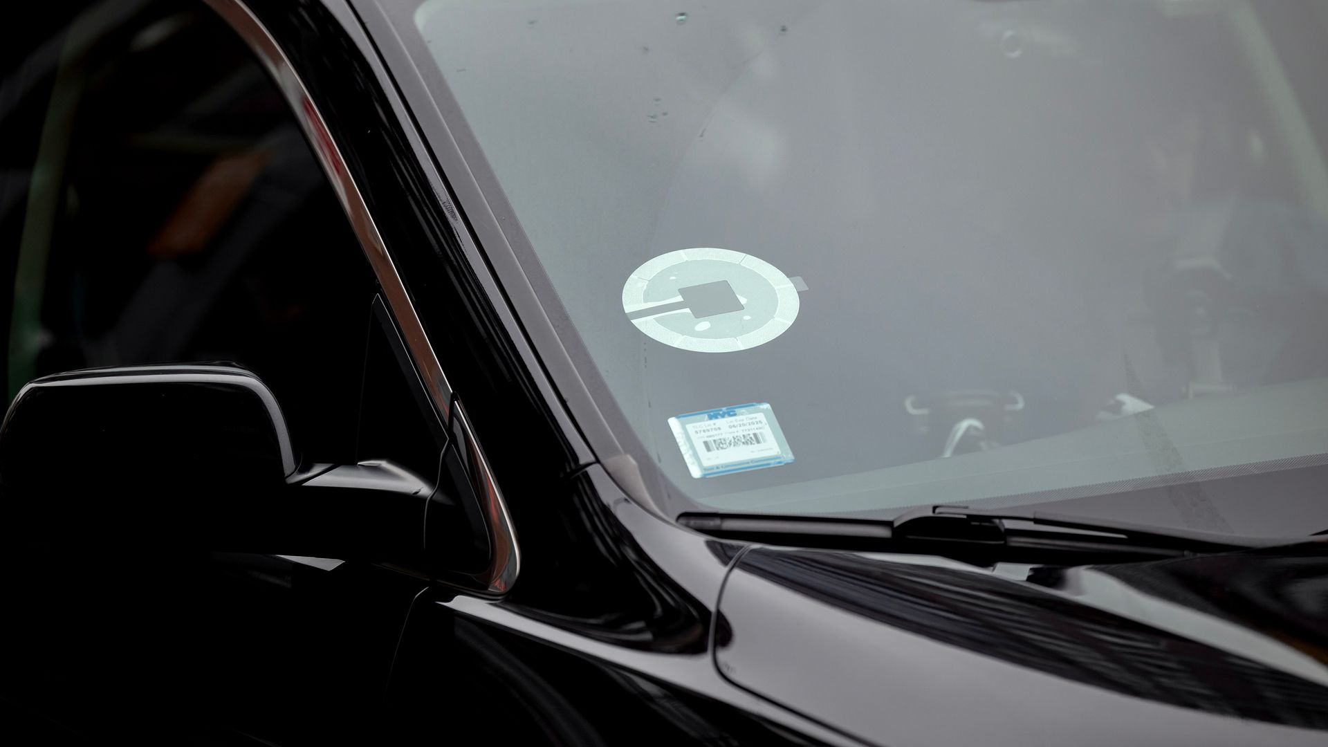Close-up of a shiny black car showing a circular white parking-permit sticker on the windshield and a blue sticker at the lower left; side mirror is visible on the left.