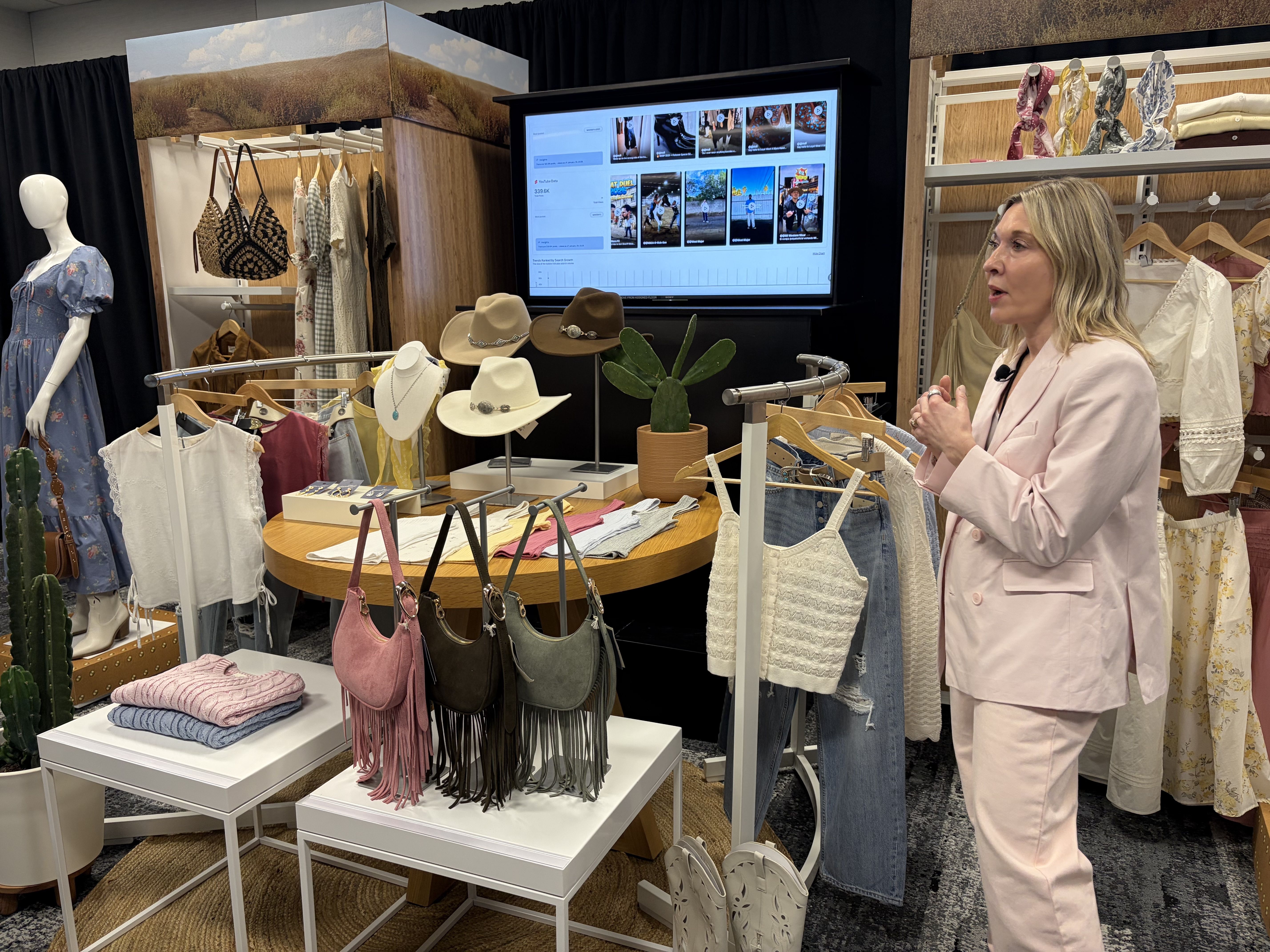 Woman in pink suit presenting a display of clothing and accessories including dresses, hats, purses, tops, jeans, and boots in a boutique-like setting with a large screen in the background.