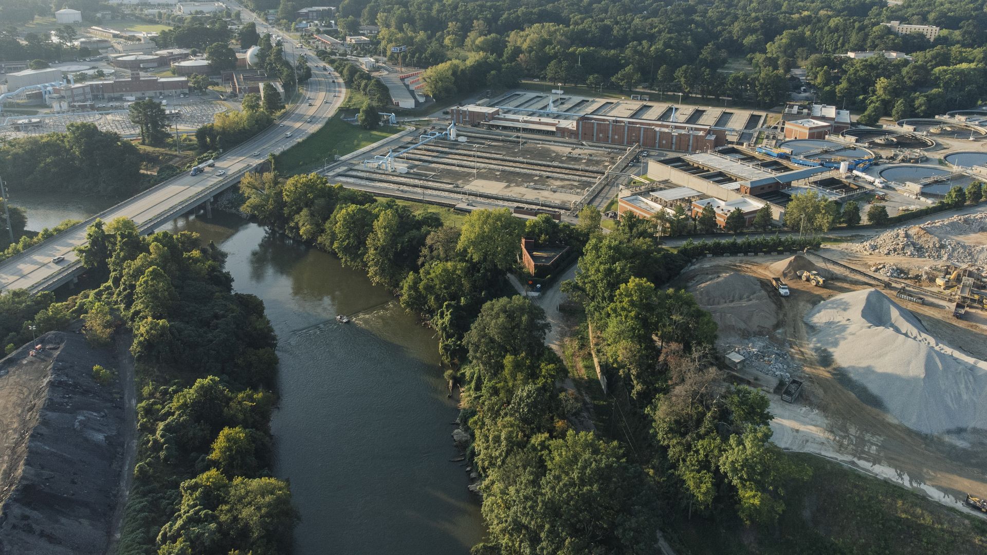 Aerial view of a water treatment facility next to a river with a highway and trees, city skyline in the hazy distance under a clear sky.