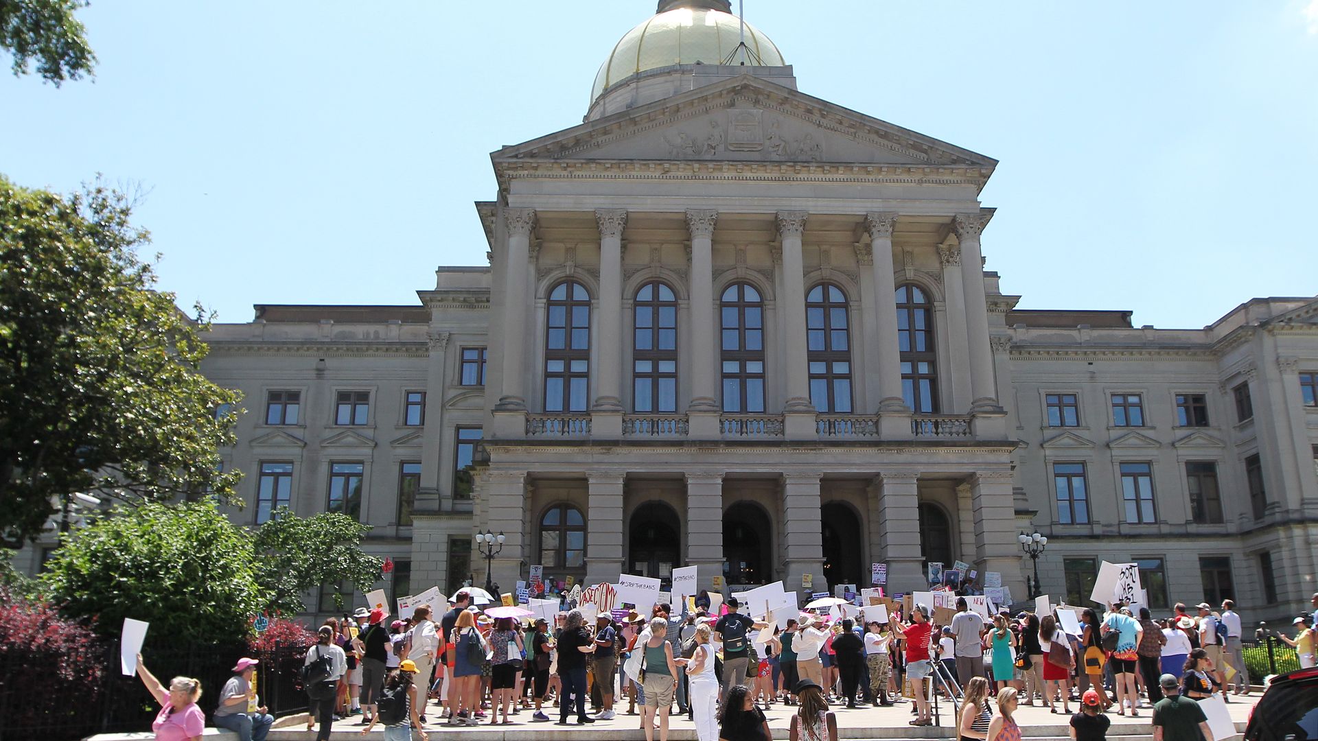 In this image, a crowd of protestors stand outside the the Georgia state capitol building. Many hold signs.