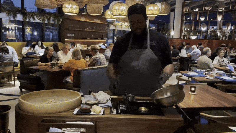 A moving image of a man cooking a pasta dish table side at a busy restaurant. He tips a cooking dish into the gas flame and the contents light on fire.