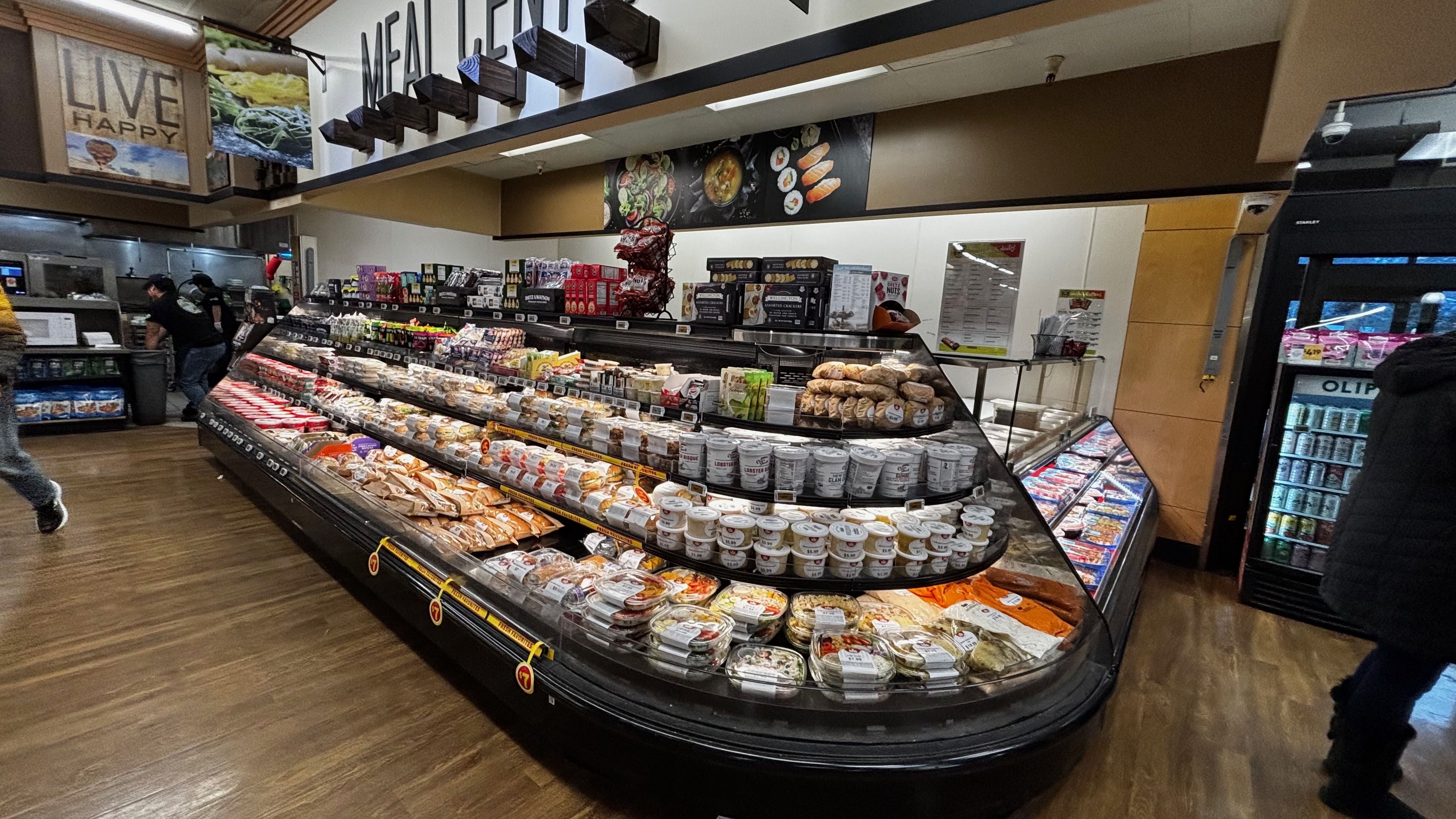 Grocery store deli section with various packaged foods, soups, and bread displayed on shelves. Brown and black decor with wooden floors and signs reading "LIVE HAPPY" and "MEAL CENTER".
