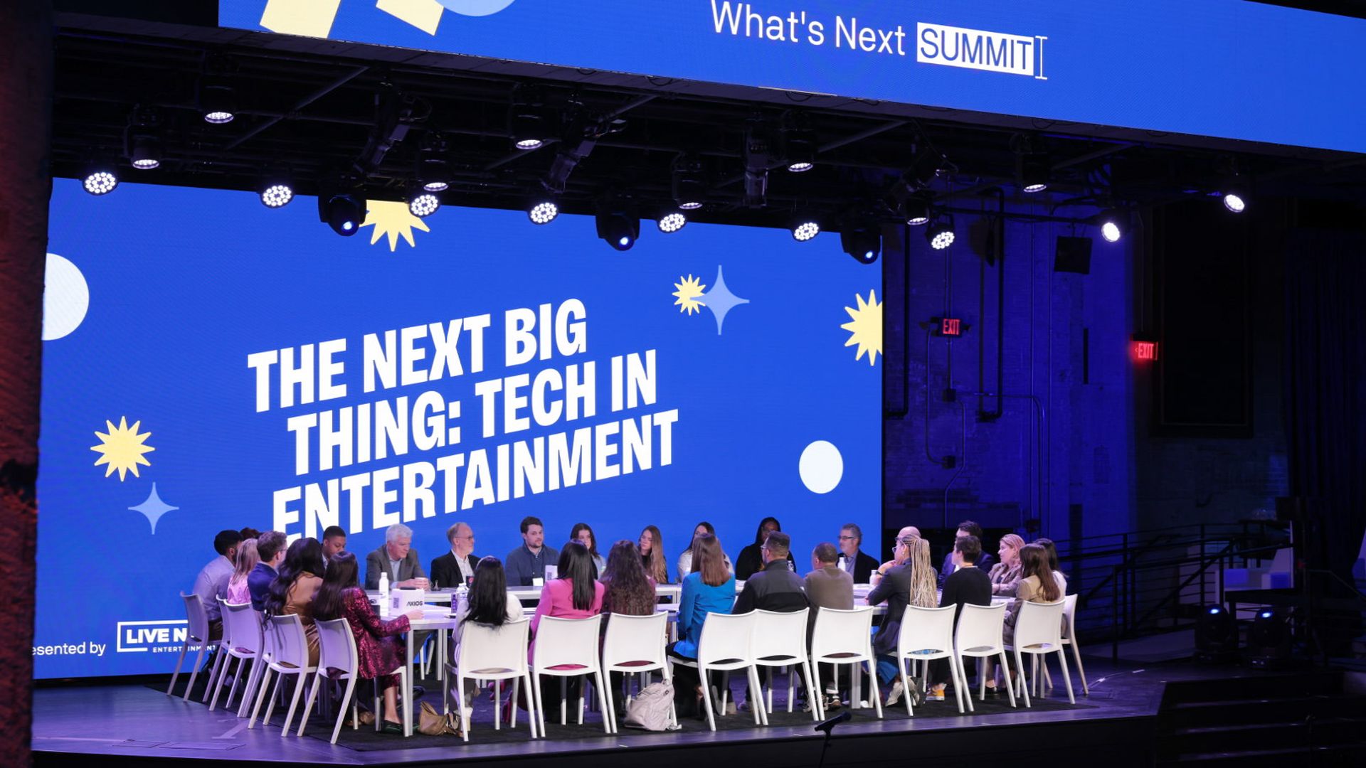About 30 people seated in white chairs around a rectangular table in front of a blue screen that reads "The Next Big Thing: Tech In Entertainment"