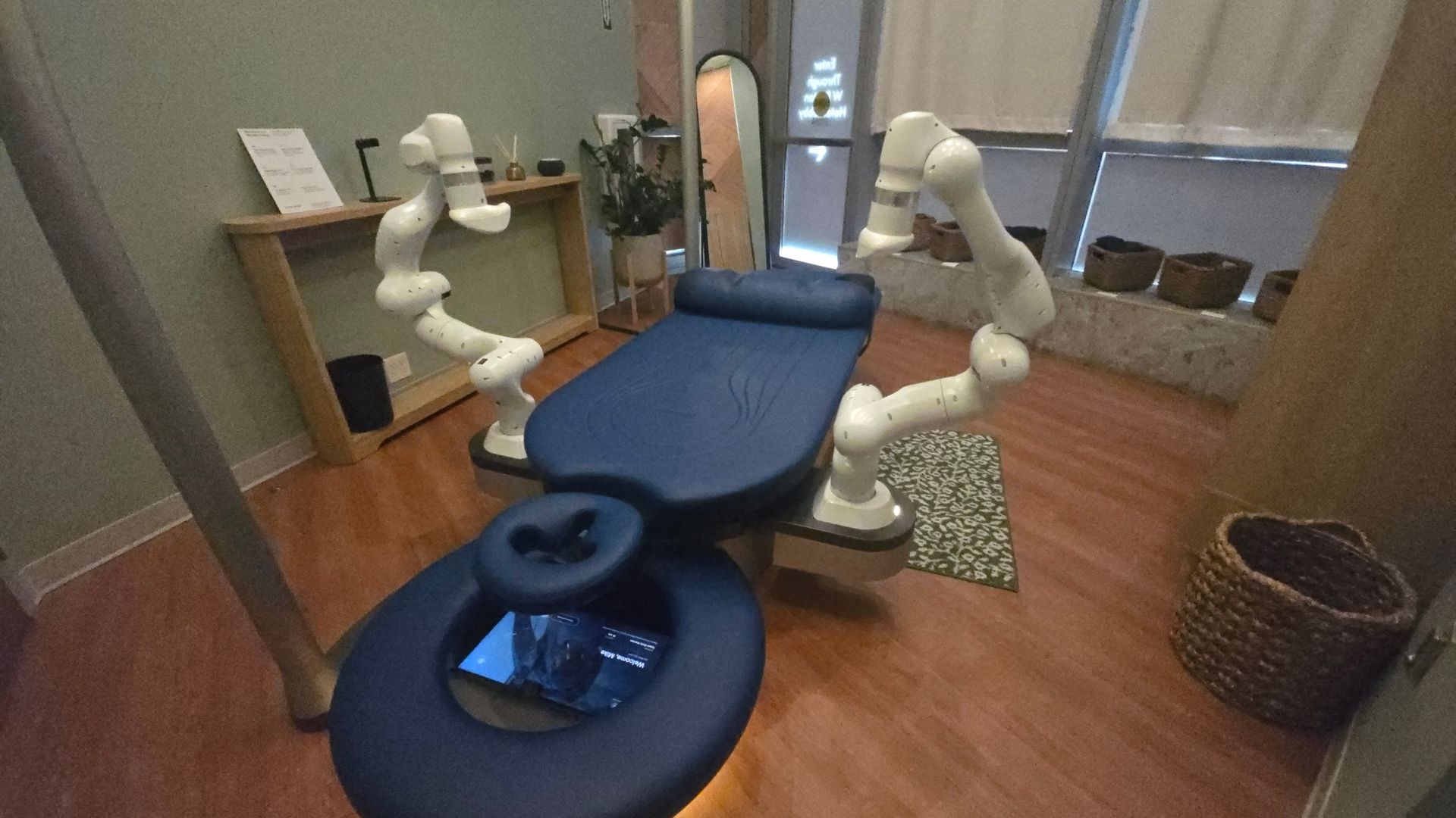 Blue padded massage chair surrounded by white robotic arms in a cozy spa-like room; wooden floor, a shelf with decor, potted plants, and a window with blinds in the background.