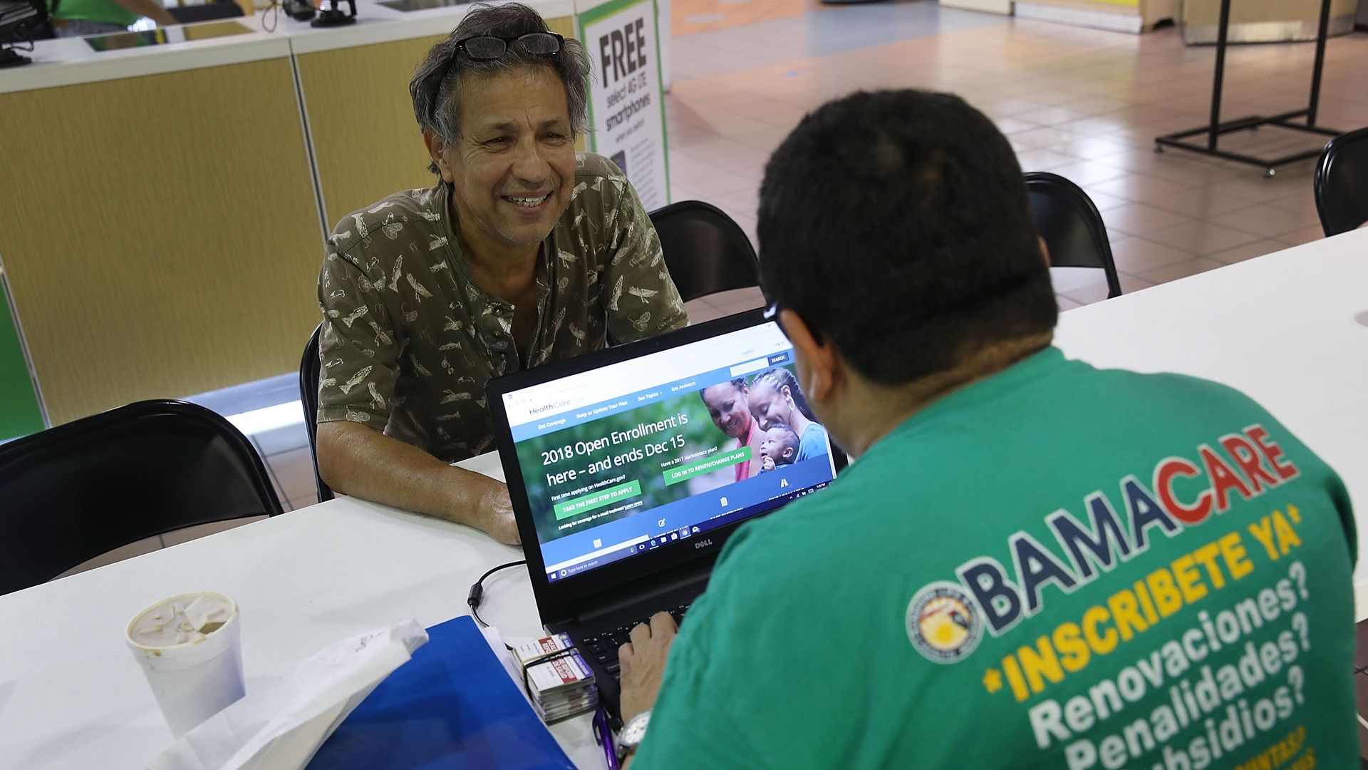 A man signs up for health coverage at a table with the help of another person at a computer.
