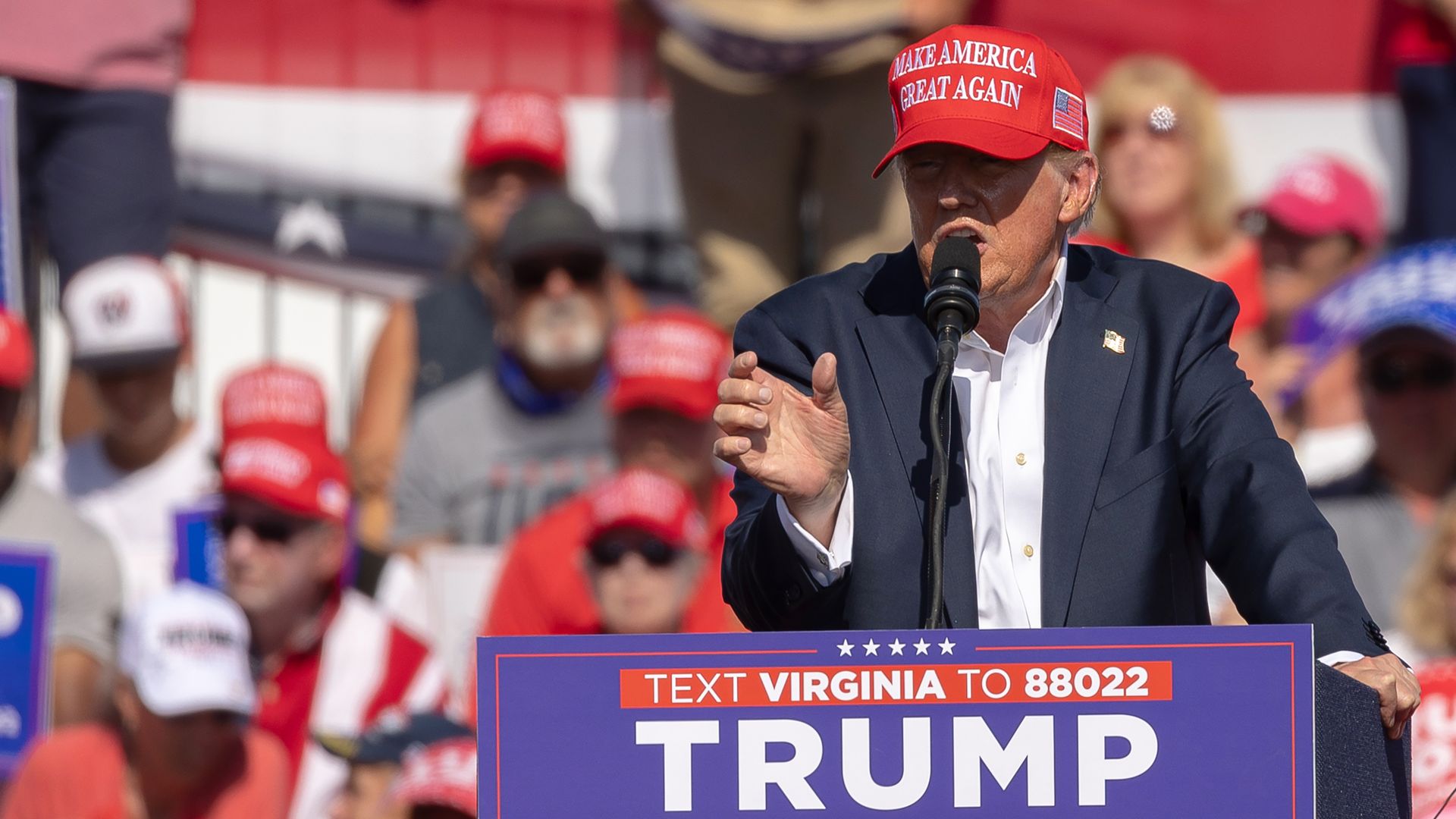 Trump, wearing a red Make America Great Again hat, dark suit and white shirt, speaks at a lectern into a microphone. A small group is visible behind him as part of a larger crowd. He's at a Virginia campaign event.