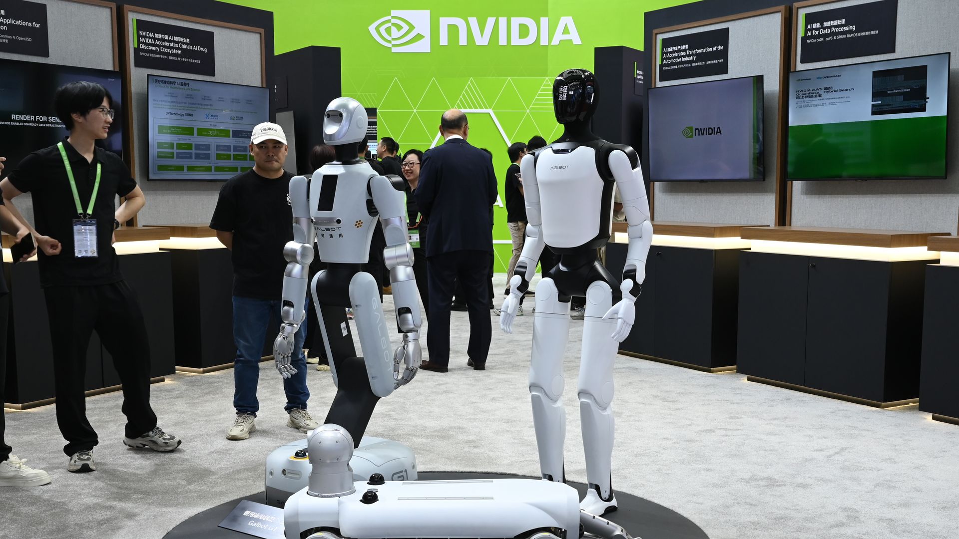 At an NVIDIA booth with a bright green wall and logo, two humanoid robots in white and black stand near a circular platform while visitors and staff discuss tech displays on screens.