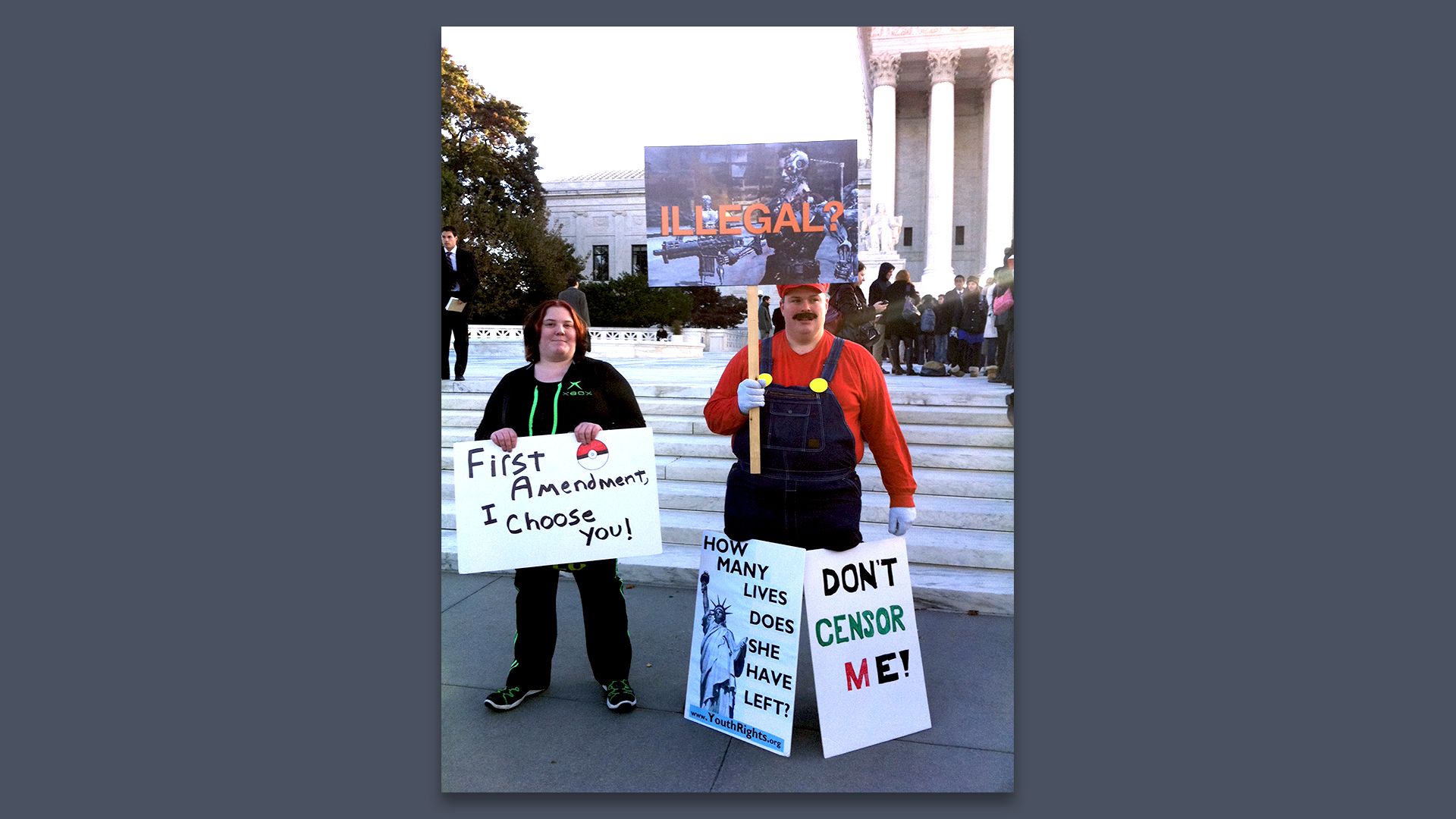 Photo of two protestors at the foot of the Supreme Court steps. One is wearing overalls and a red shirt. Another holds a sign that says: "First Amendment, I choose you."