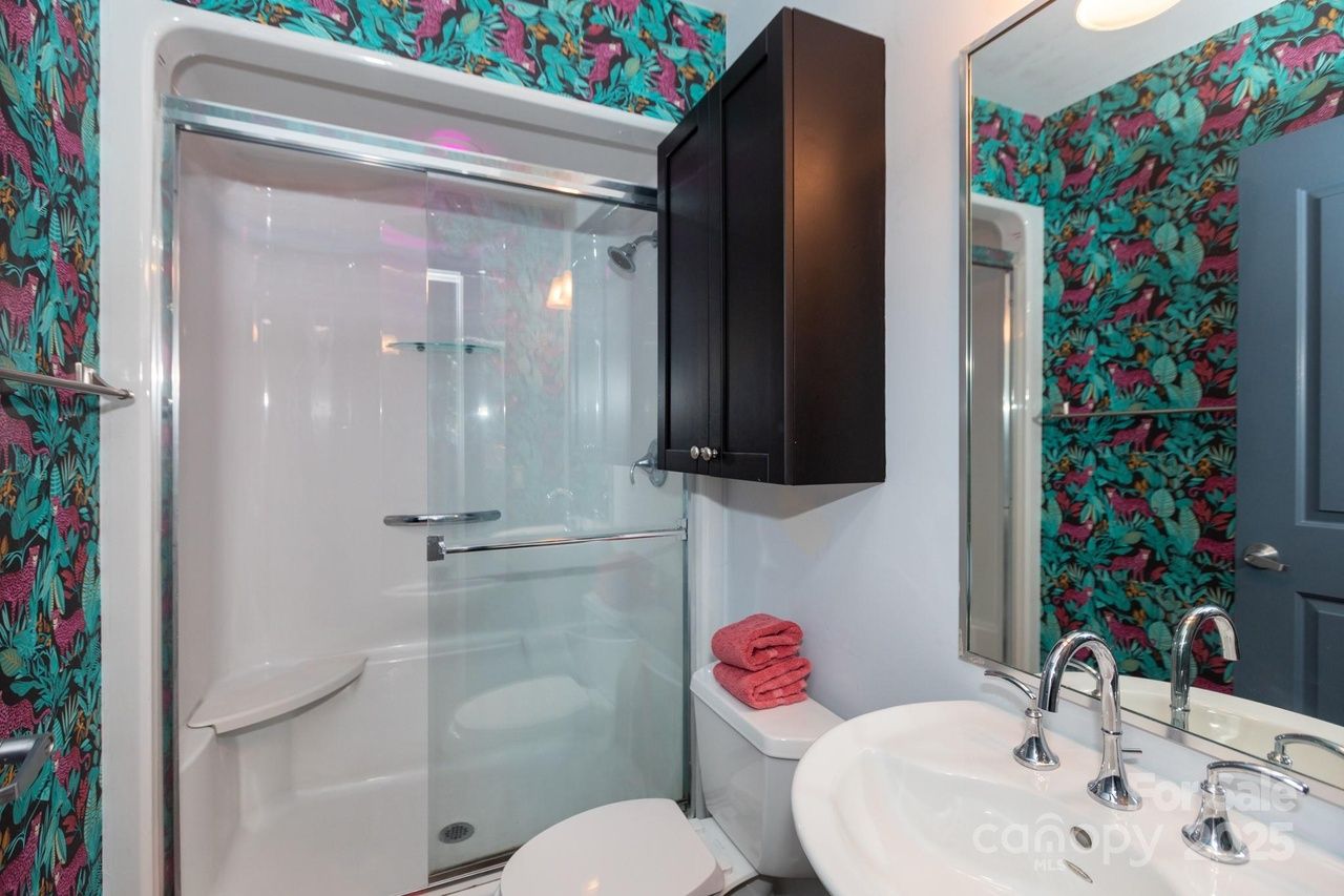 Bathroom with white sink, chrome faucet, toilet with red towels, black wall cabinet, glass shower doors, and vibrant wallpaper with teal leaves and pink animals.