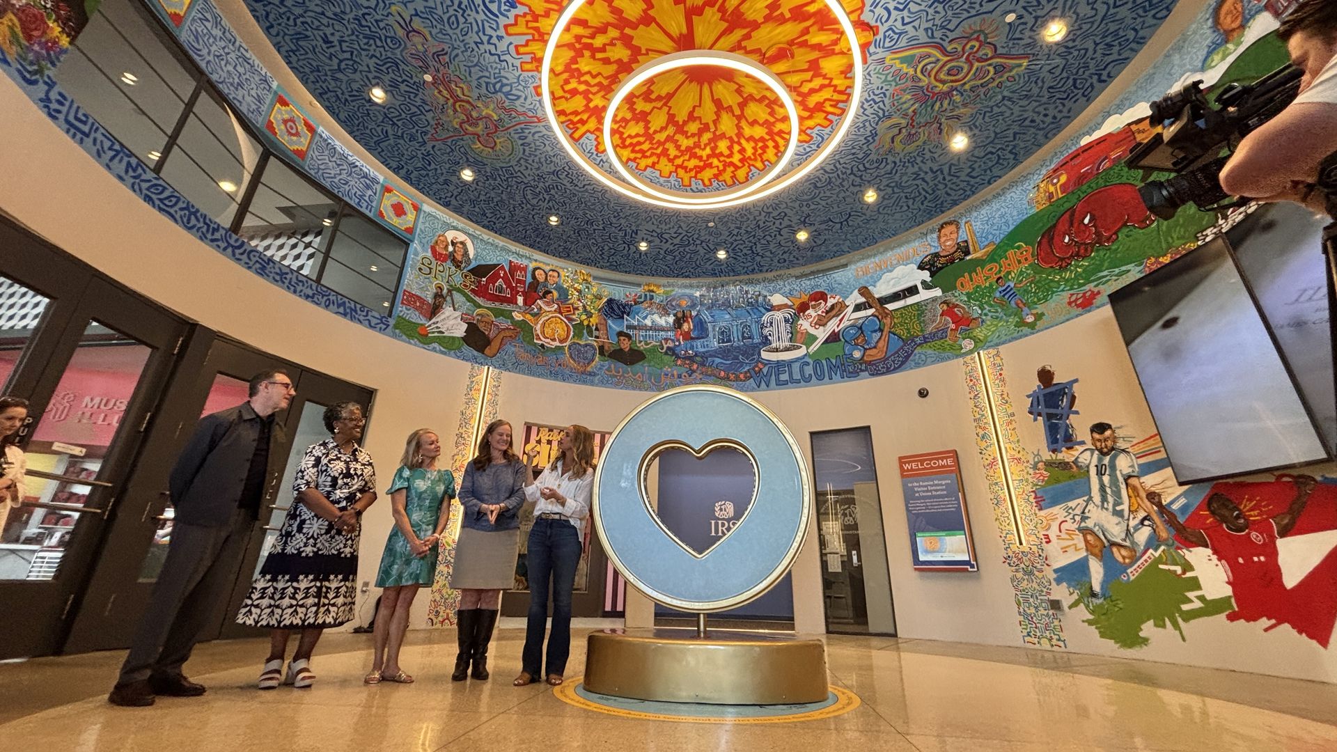 A curved mural lobby with a bright orange sun chandelier overhead. In the center, a blue circular heart sculpture on a pedestal; six people stand along the wall while a photographer films.