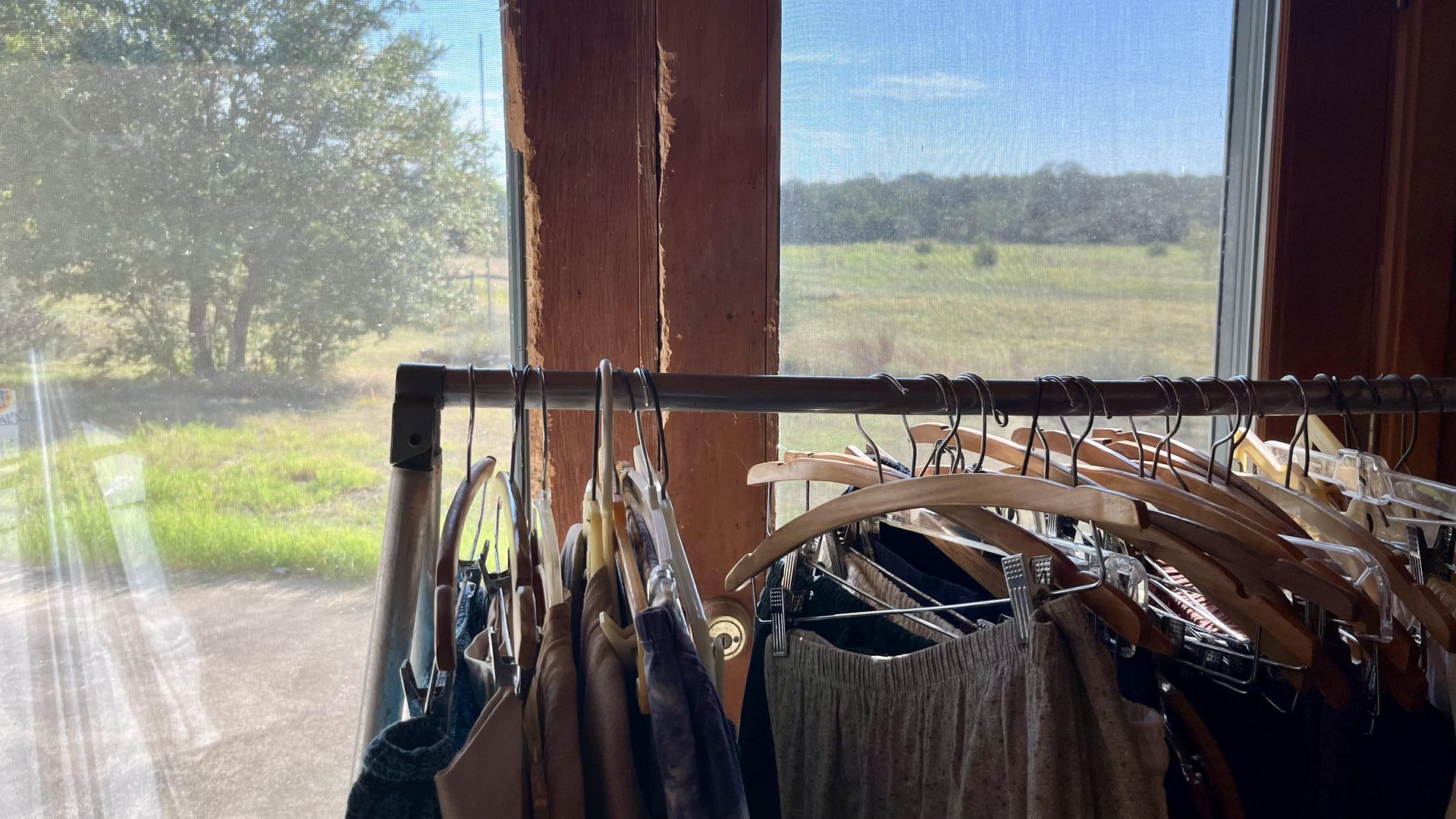 Clothing hangs on a rack in front of a window showing a bright, sunny landscape in the Texas Hill Country with trees and grass.