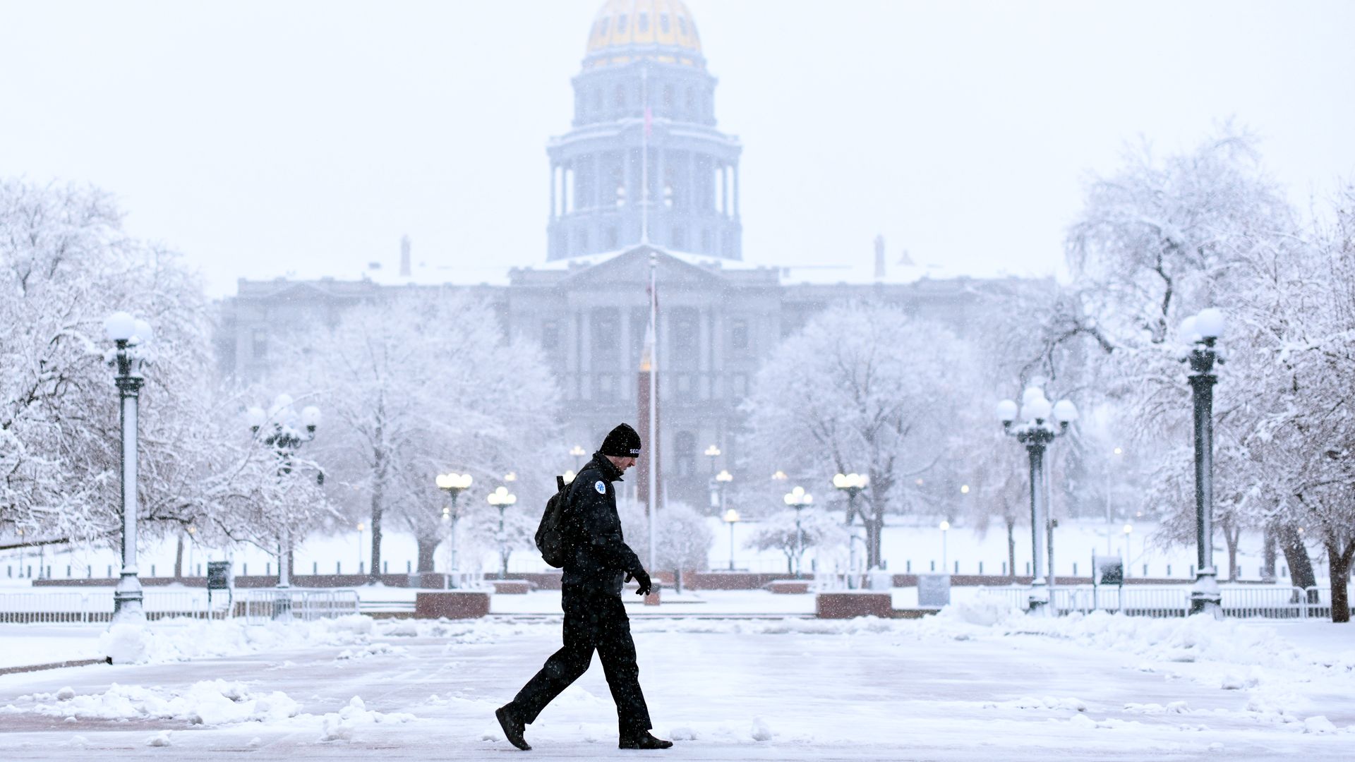 A man walks along a snowy street across from the Colorado State Capitol, which is grey with a golden dome.