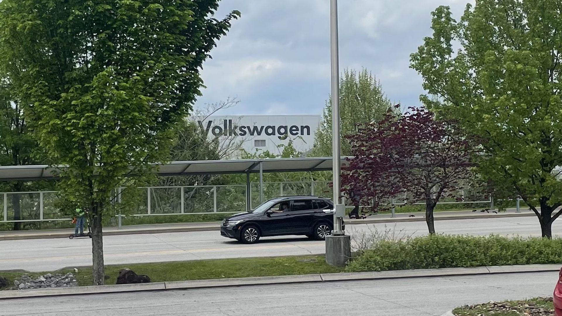 A car drives down a quiet street with a Volkswagon plant in the distance behind a row of trees.