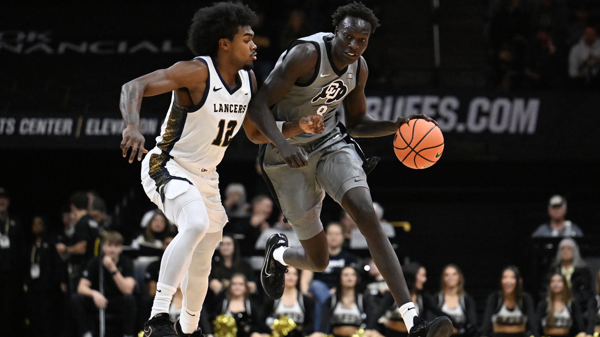 Two basketball players in action; Bangot Dak in gray CU Buffs jersey with ball tries to advance while a player in white Lancers jersey defends. Cheerleaders and audience blurred in background.