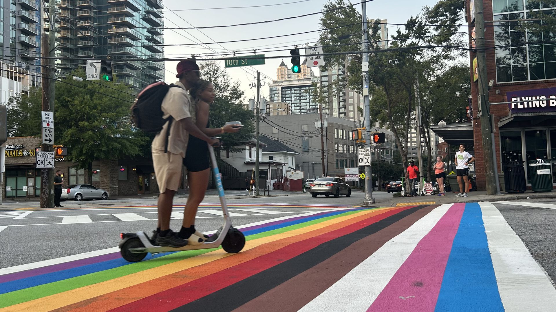 A photo of two people riding a scooter over a rainbow crosswalk